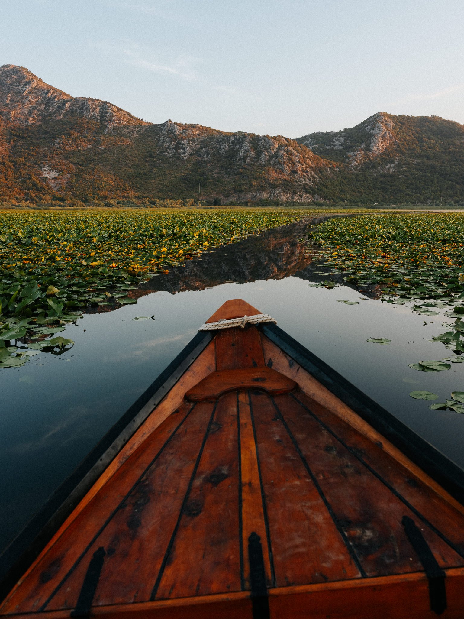 View from a canoe on a calm river with lily pads, reflecting surrounding mountains and a clear sky.