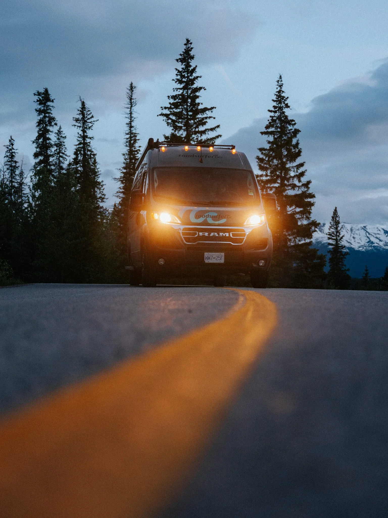 A camper van on a mountain road with tall trees in the background during dusk or dawn.