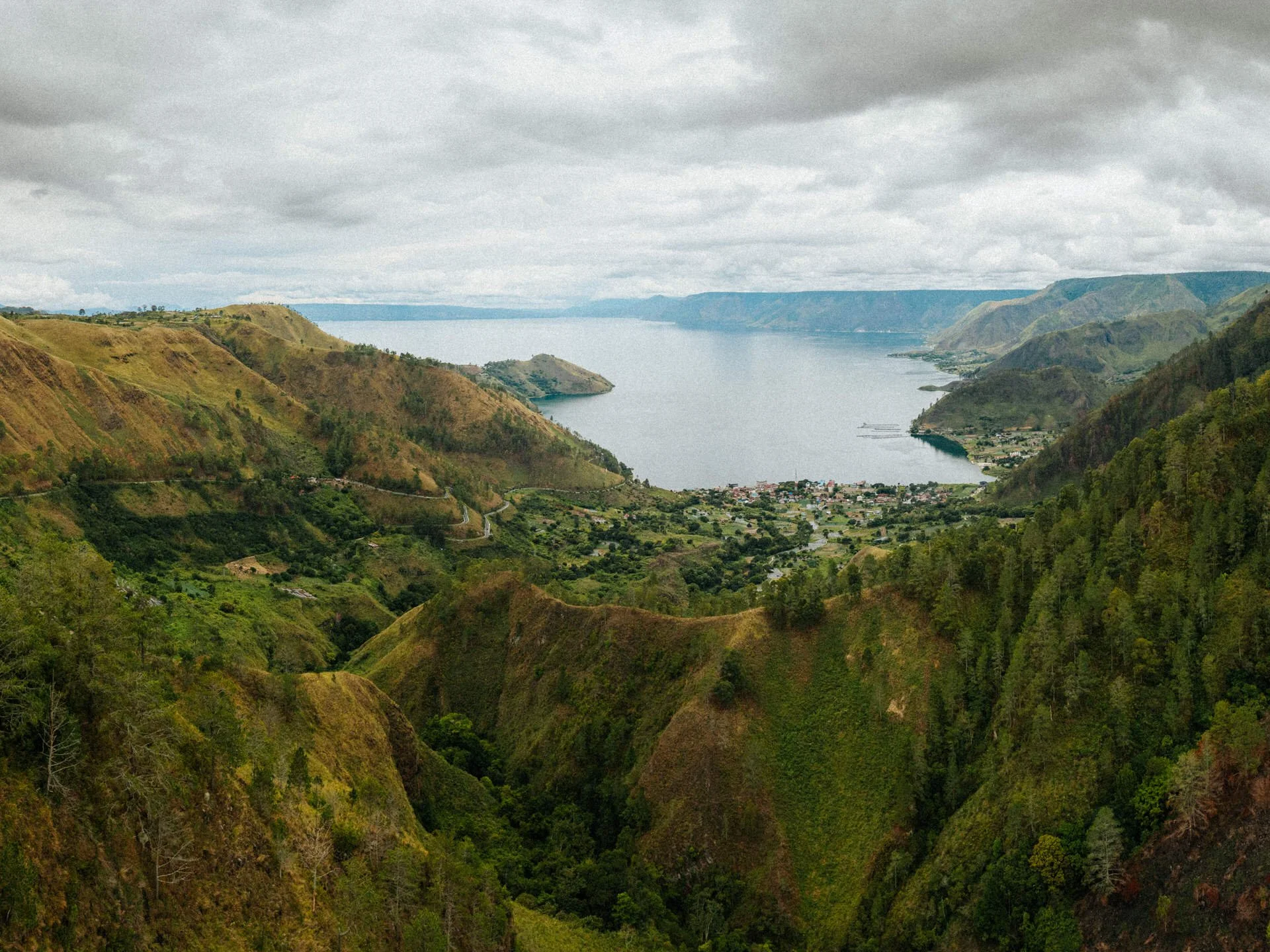 A panoramic view of a lake surrounded by green hills and mountains under a cloudy sky.