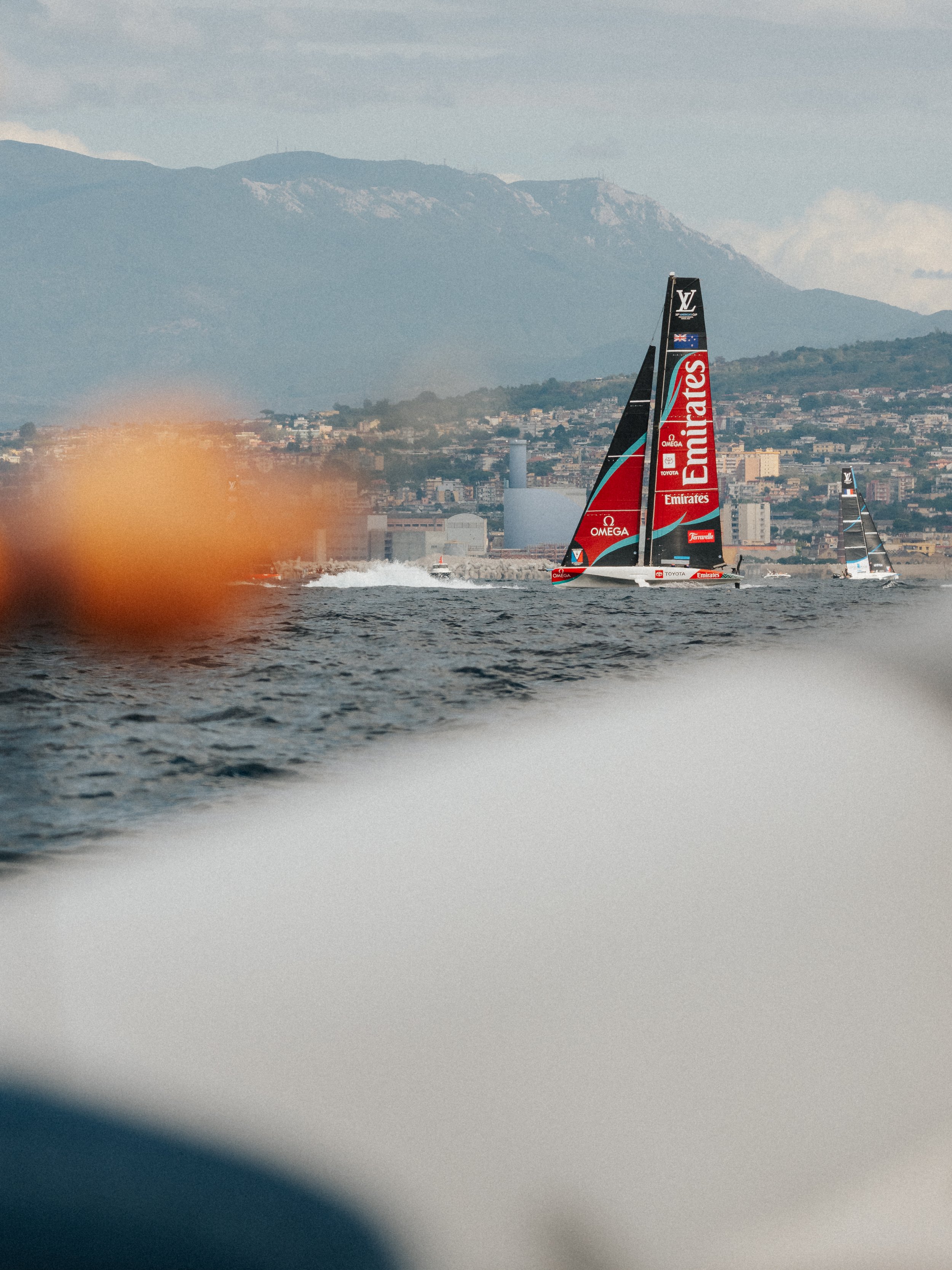 Sailboats racing on water with cityscape and mountains in the background, one sailboat prominently displaying sponsors and logos.