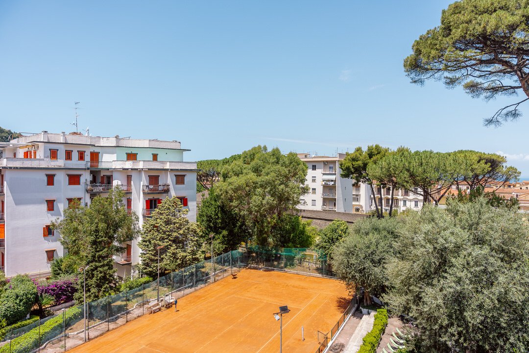 A red clay tennis court surrounded by green trees and residential buildings, with a clear blue sky overhead.