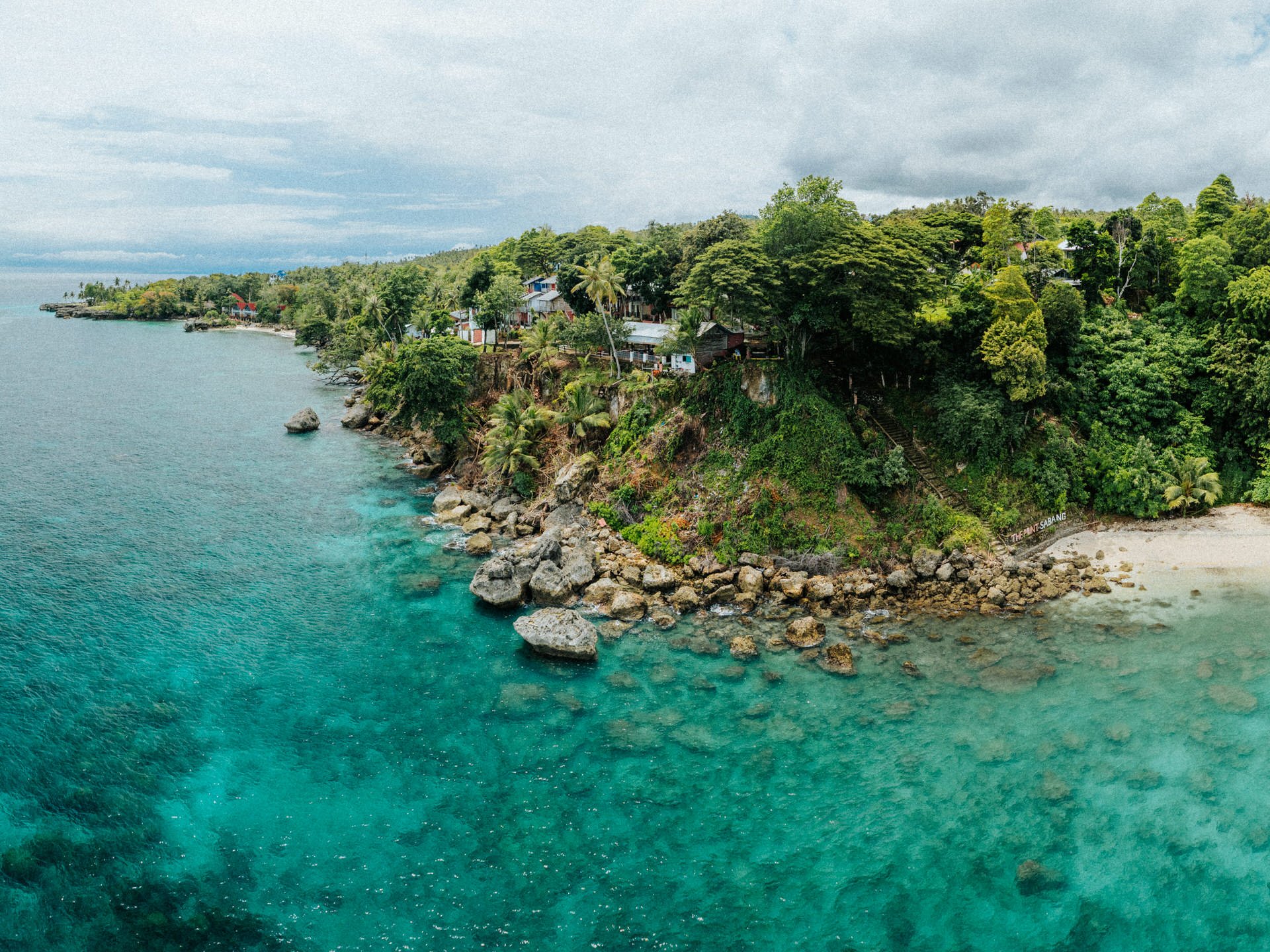 A coastal scene with turquoise water, rocky shoreline, and lush green trees on a hillside with houses.