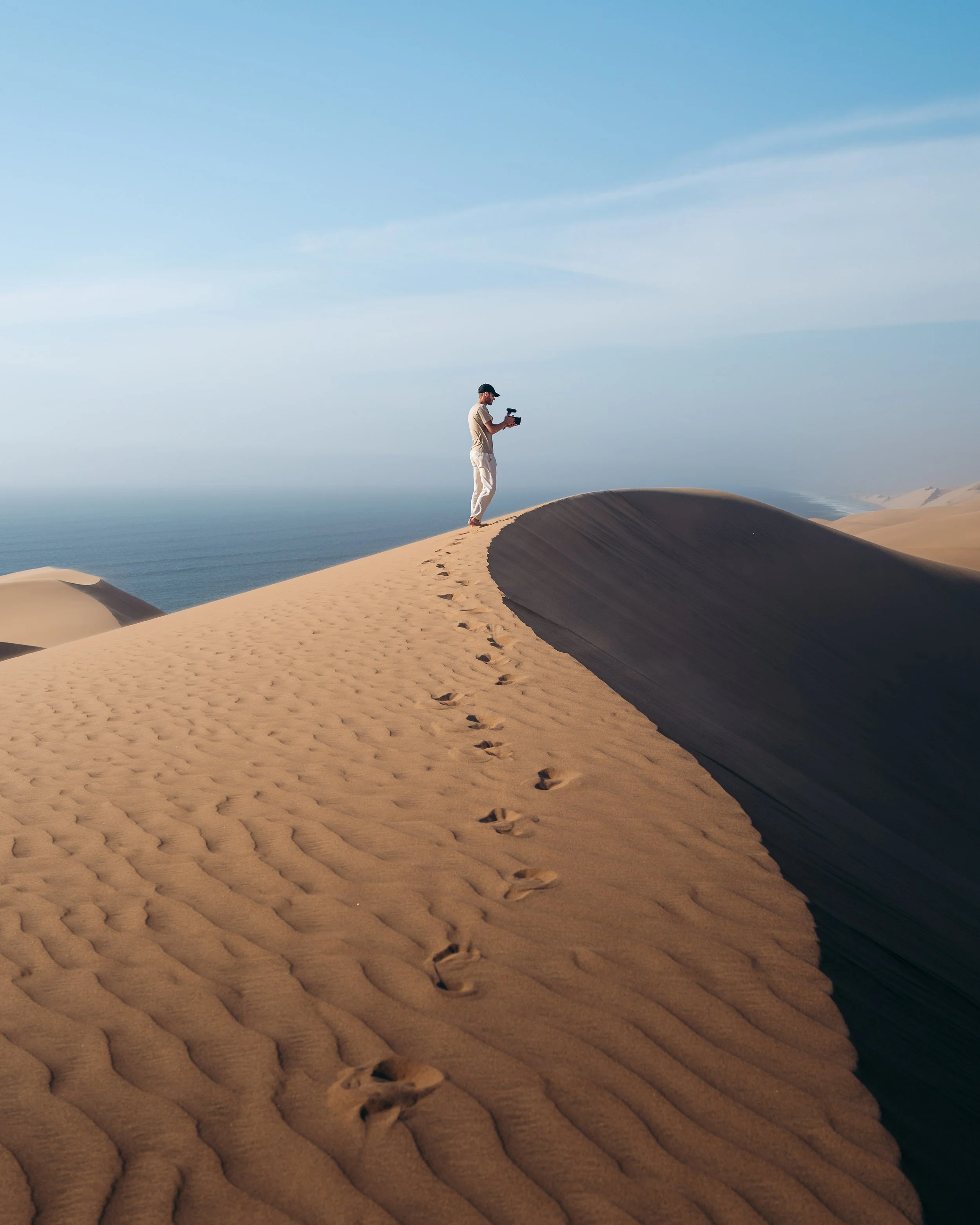A person standing on a sand dune taking a photo with a camera, with footprints leading up to them on the sand dune, overlooking the ocean under a blue sky.