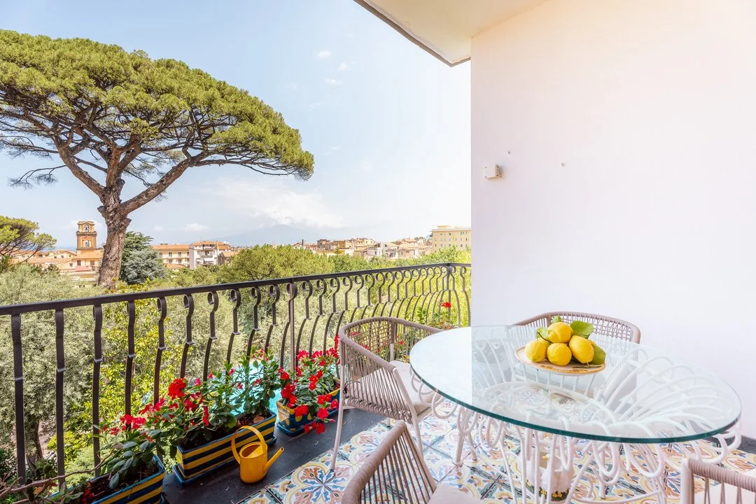 Balcony with a round glass table, white chairs, and a bowl of lemons, overlooking trees and buildings with a large pine tree in the background.