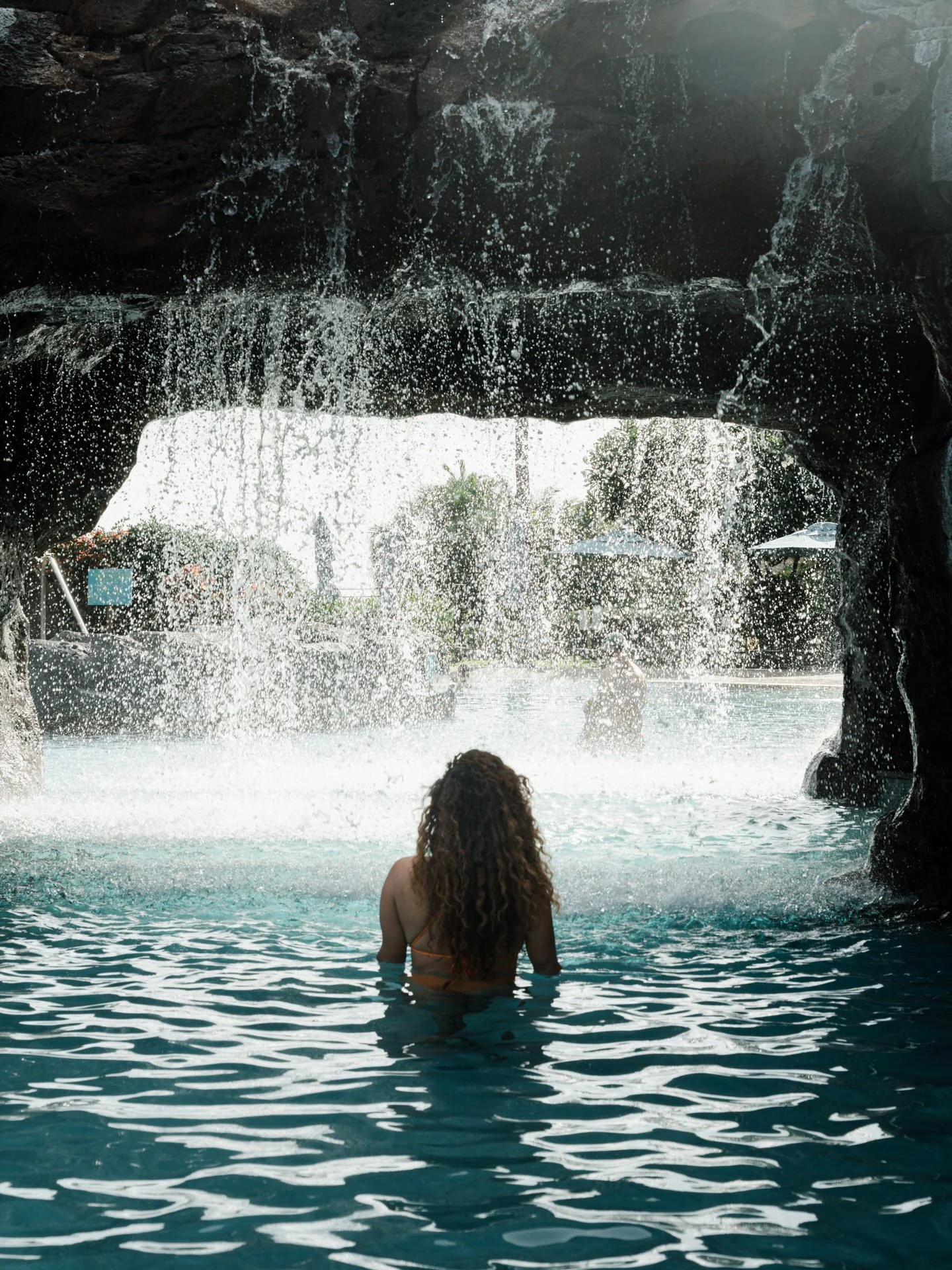 Woman with curly hair swimming in a pool under a waterfall cave opening.