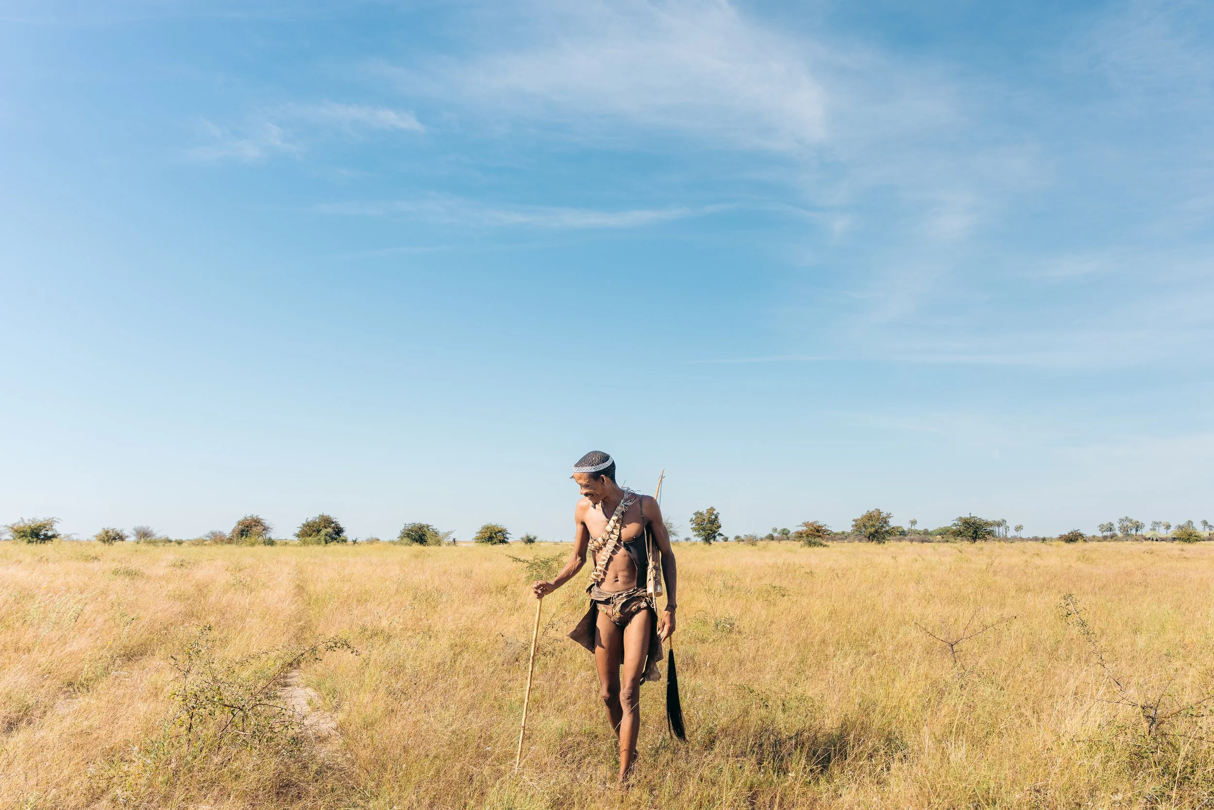 A person with traditional attire walking in a grassy plain under a blue sky with some clouds, and sparse trees in the background.