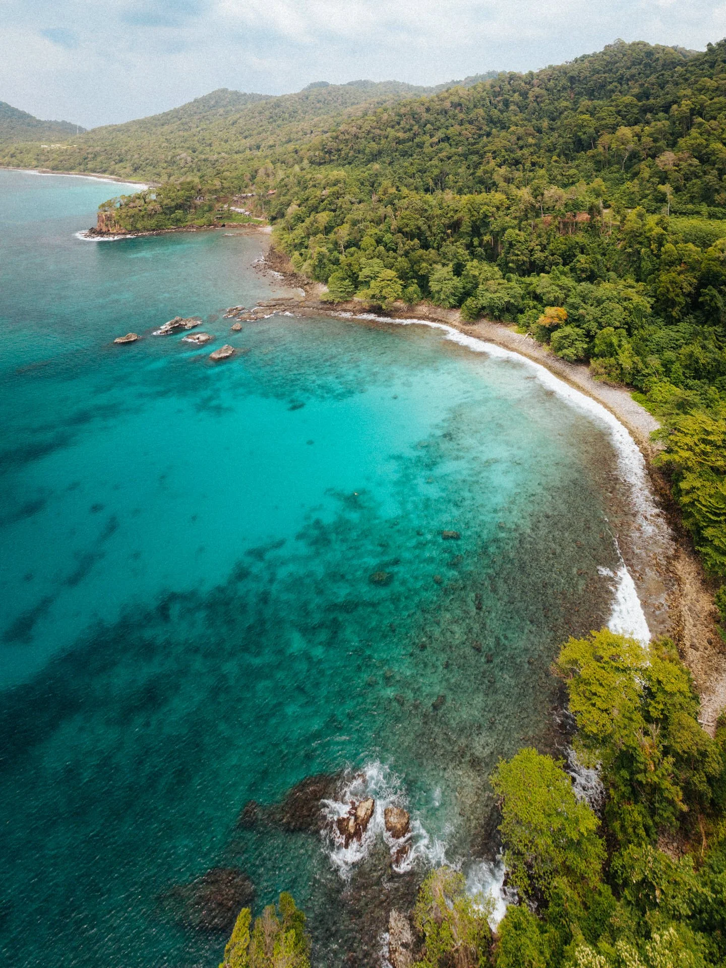 Aerial view of a tropical coastline with clear turquoise water, lush green forested hills, and rocks along the shore.