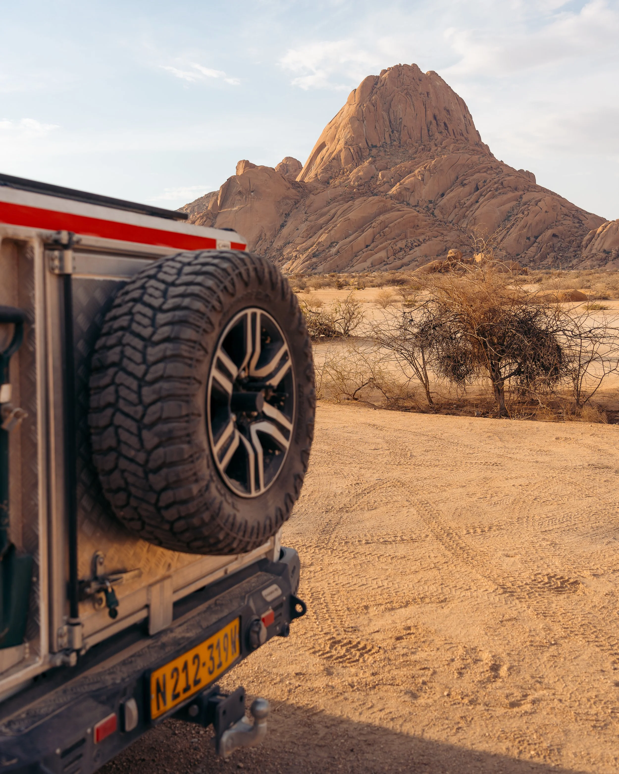 A desert landscape with large rocky mountains in the background, dry bushes, and a vehicle with a spare tire on the back in the foreground.