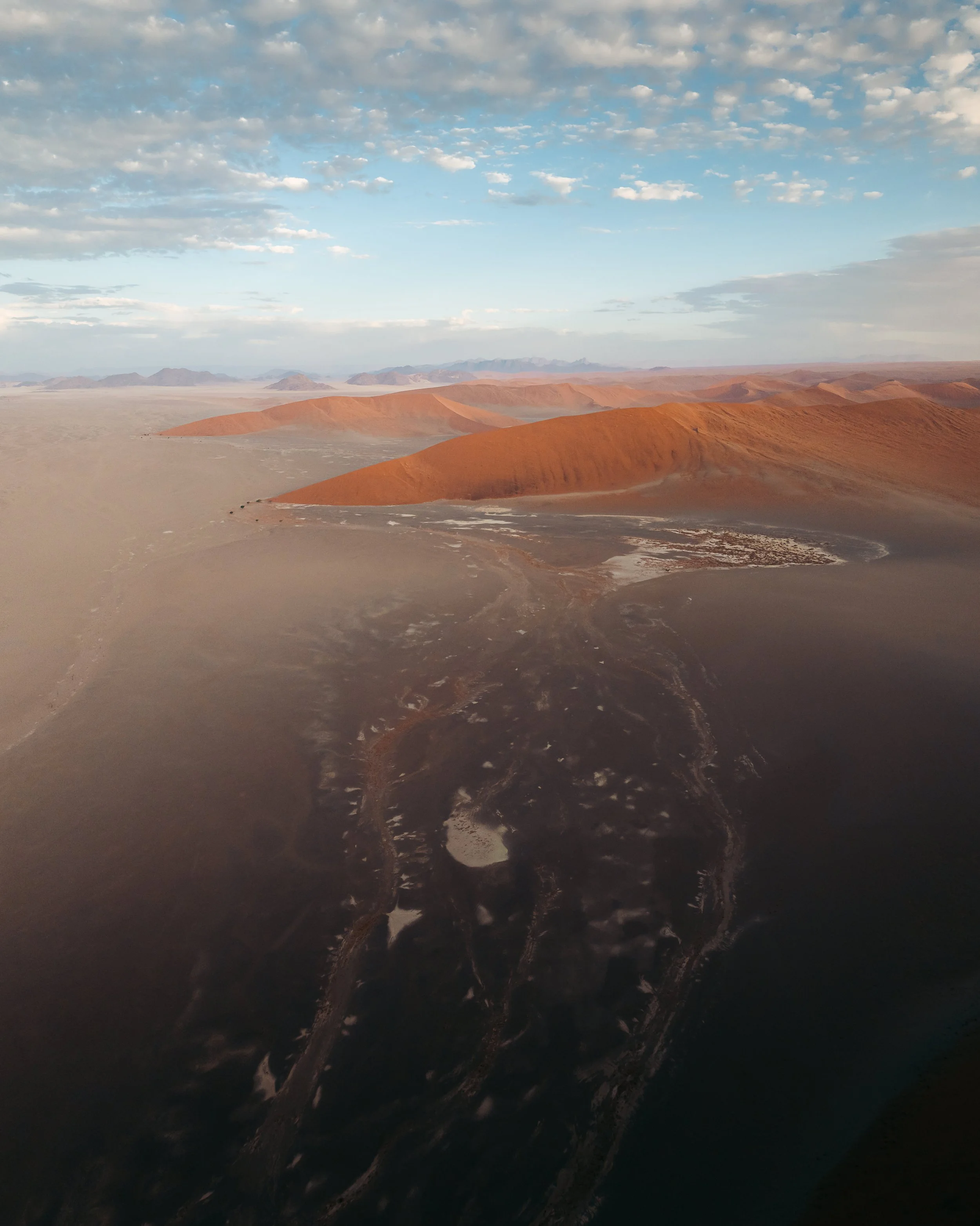Aerial view of desert mountains with reddish-orange sand dunes, some water bodies, and a sky with scattered clouds at sunset or sunrise.