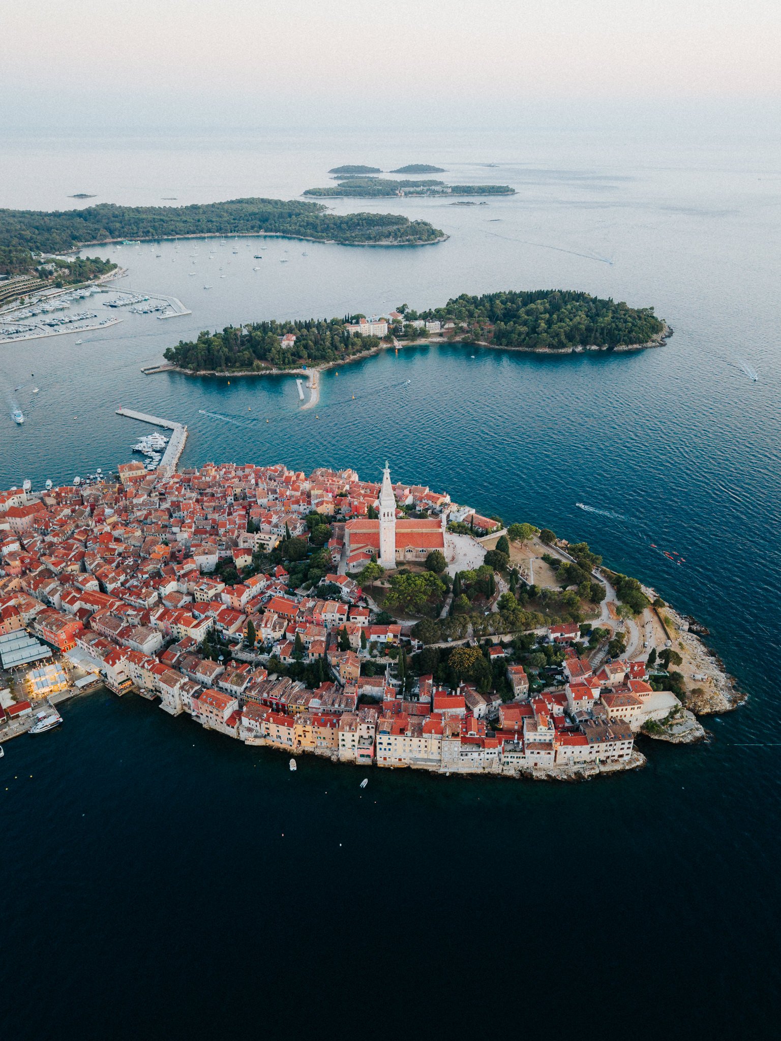Aerial view of a historic coastal town with red-tiled roofs, a prominent church tower, surrounding stone walls, and a marina with boats, on a small peninsula extending into the dark blue sea.