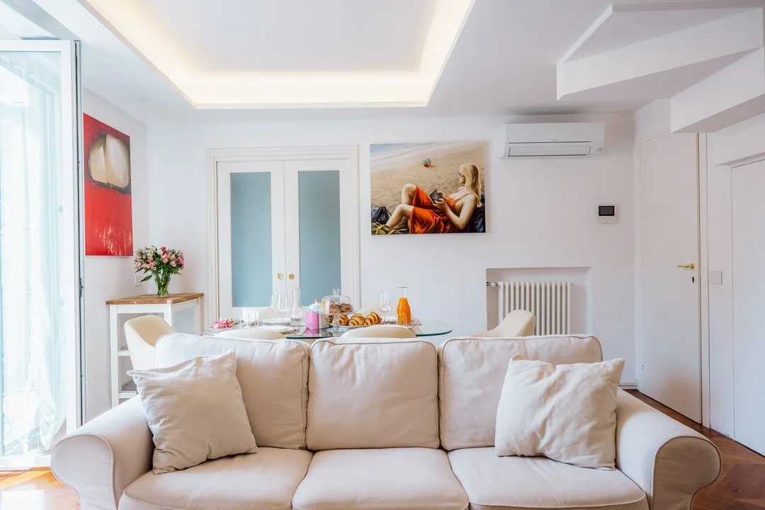 Bright, modern living room with white walls, a beige sofa with matching pillows, a dining table set with breakfast items and drinks, a wall-mounted air conditioner, artwork including a portrait of a woman on a beach, and double glass doors leading ou