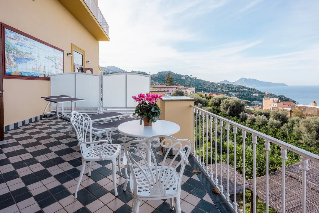 Balcony with a table and chairs overlooking a hilly landscape and the ocean under a partly cloudy sky.