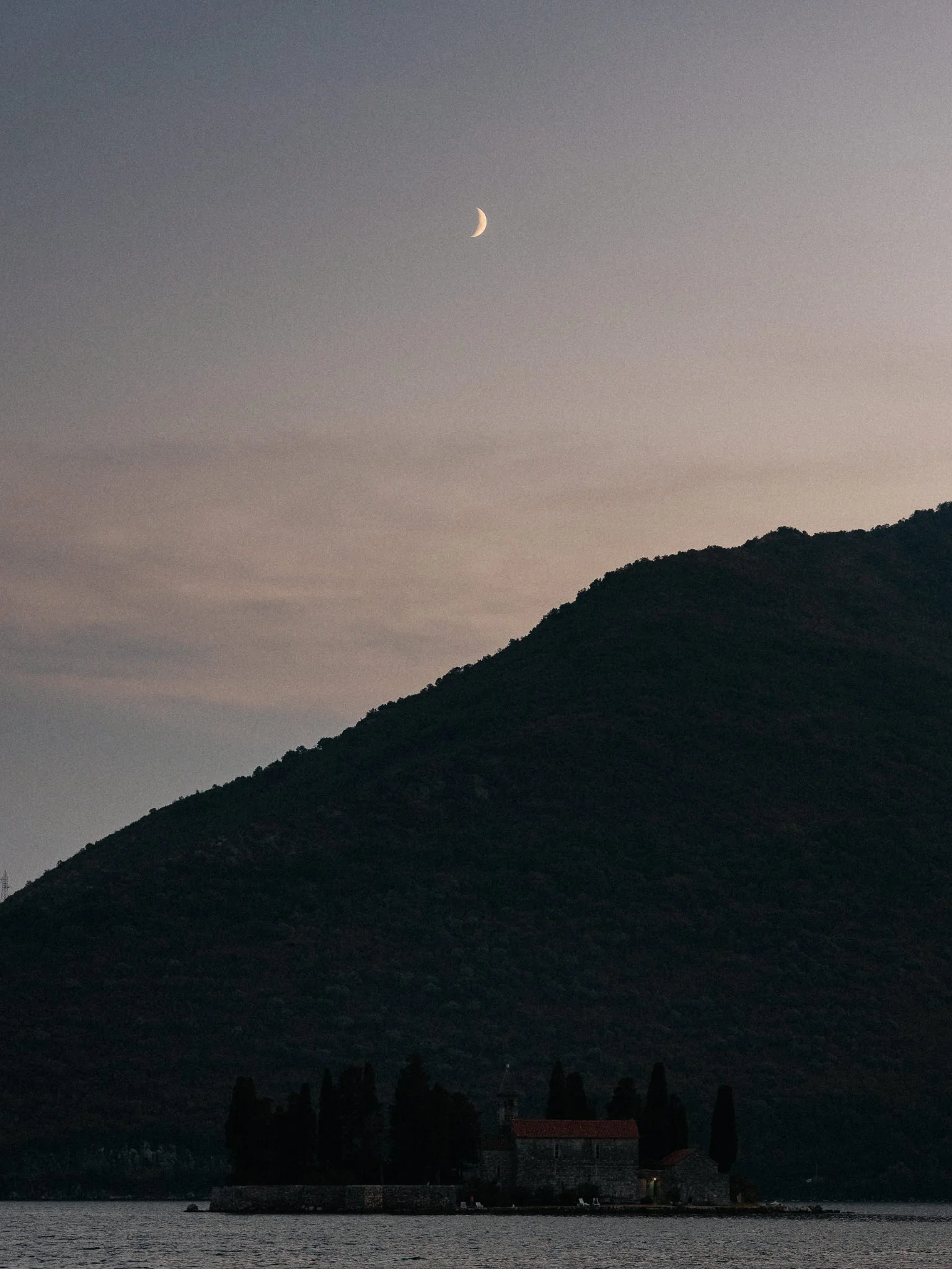 Nighttime scene of a lake with a historic stone building on water and mountains in the background, with a crescent moon in the sky.