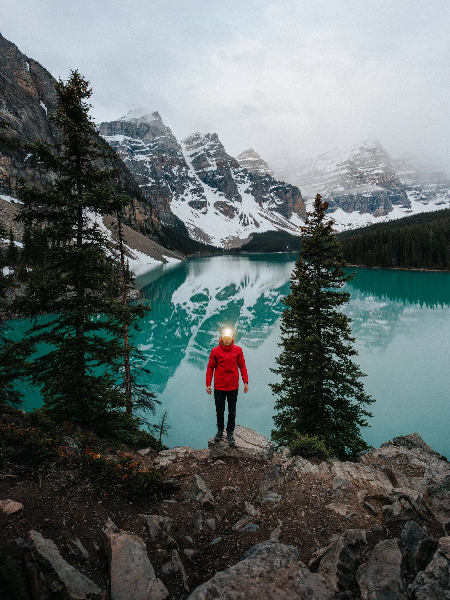 A person in a red jacket standing on rocks at the edge of a lake surrounded by snow-capped mountains and pine trees.