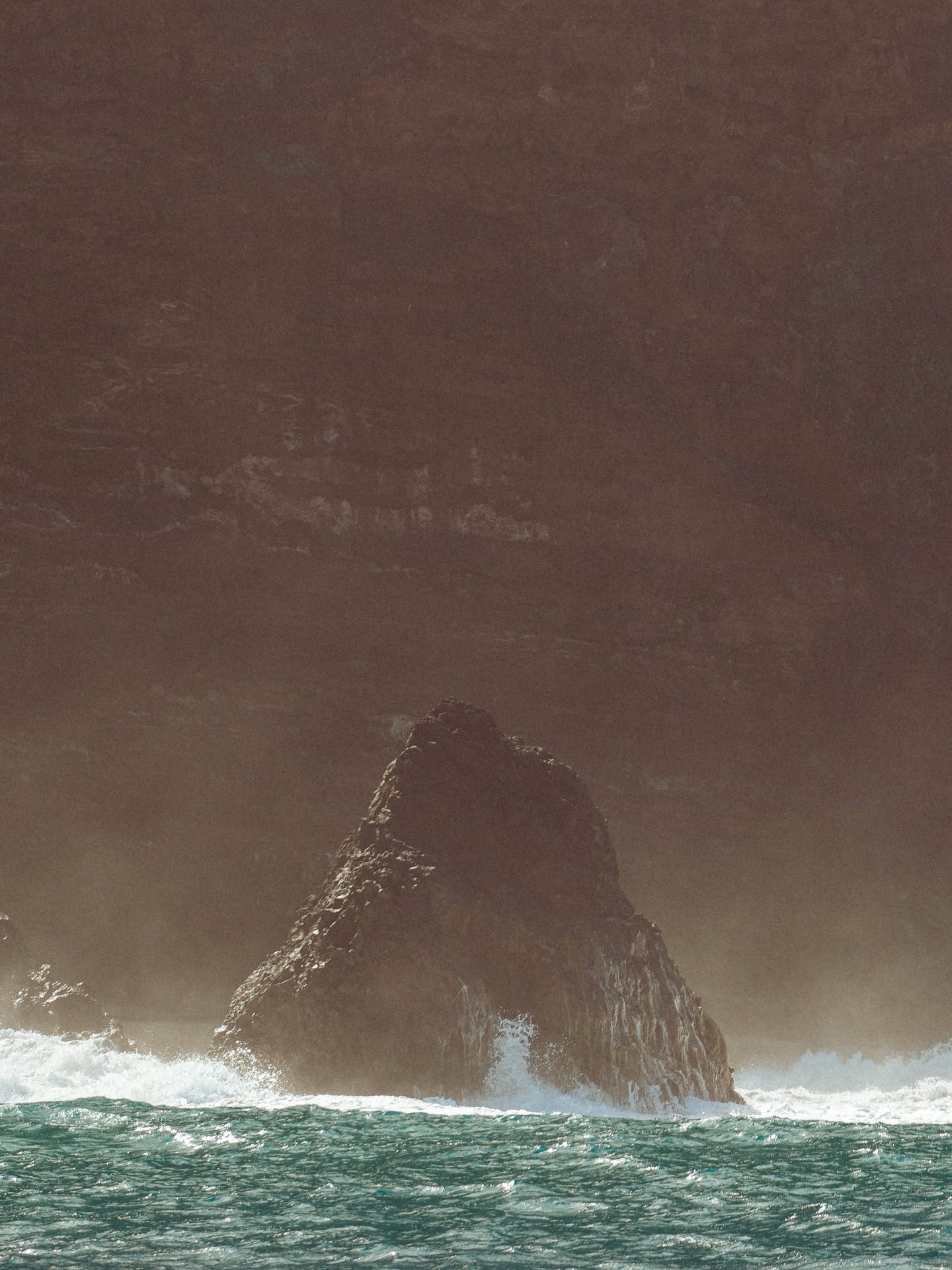 A large dark rocky island or sea stack with waves crashing at its base, under a cloudy sky.