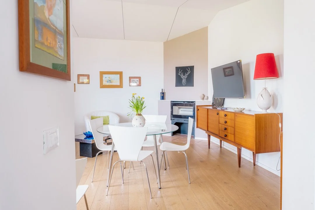 Living room with a round dining table, white chairs, a white couch with green cushions, a television on a wooden sideboard, red lamp, and wall art.