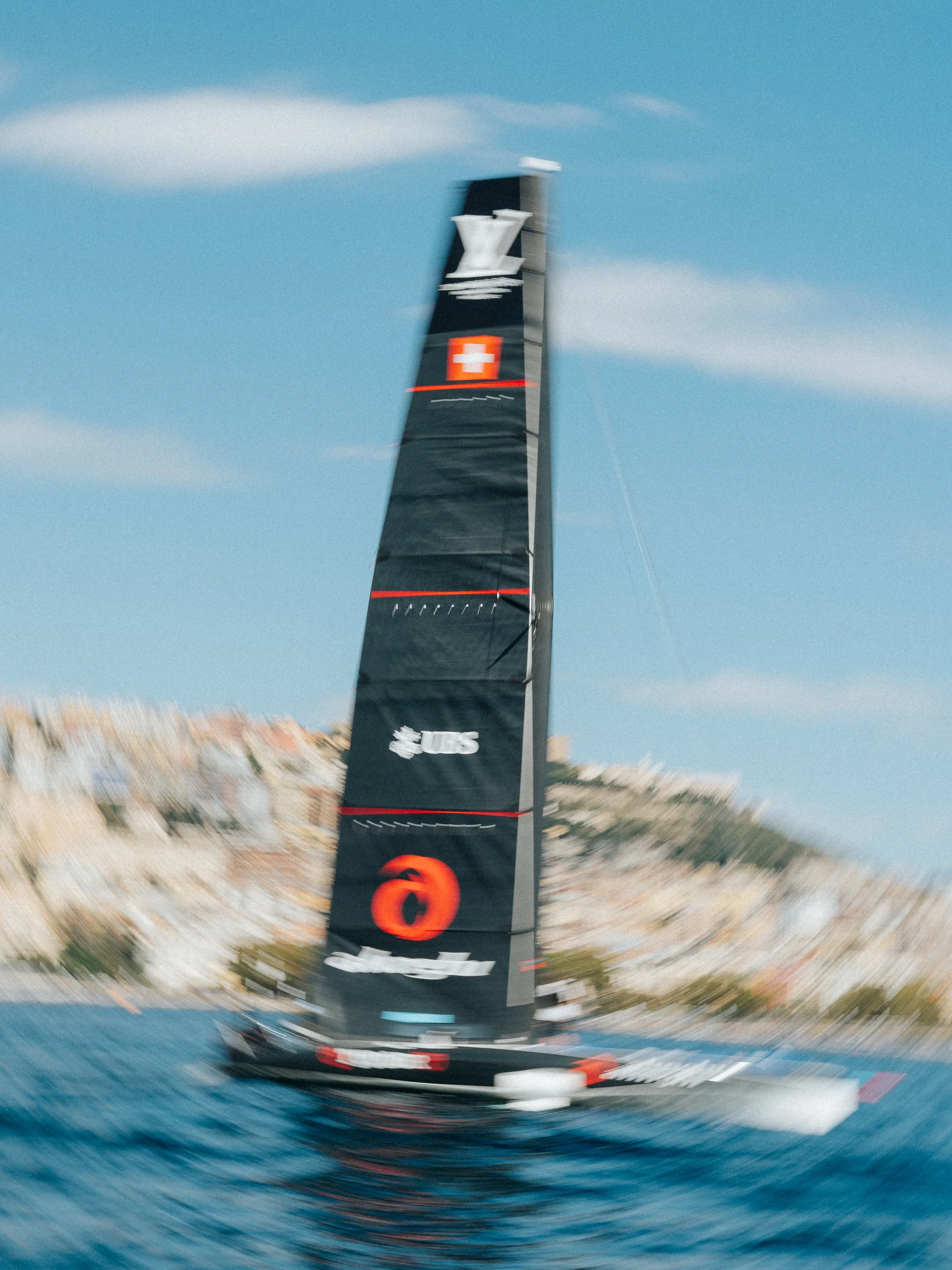 A tall black racing sailboat with a prominent sail featuring red and white branding, speeding through blue water under a clear sky with wispy clouds.