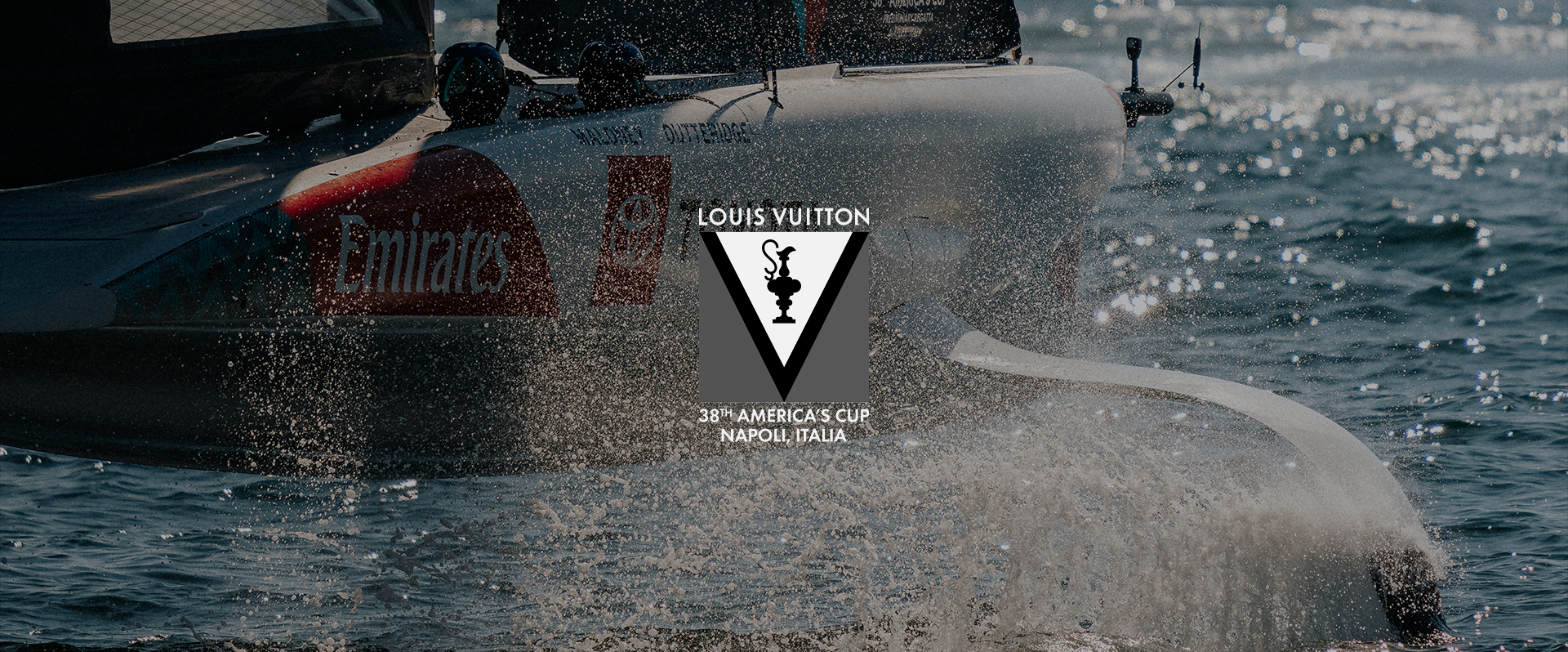 A sailboat speeding through the water during the Louis Vuitton America's Cup event in Naples, Italy, with logos of Emirates and Toyota visible on the boat, and water splashing around.