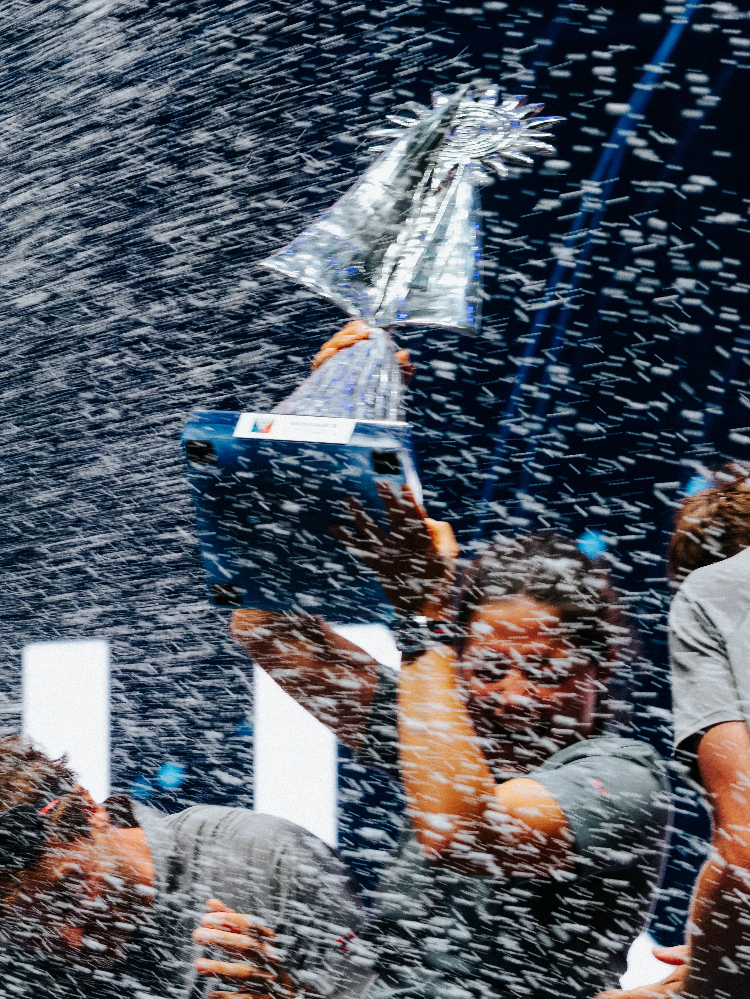 A person is holding a shiny, silver trophy with a sunburst design on top, surrounded by confetti or snow-like particles.