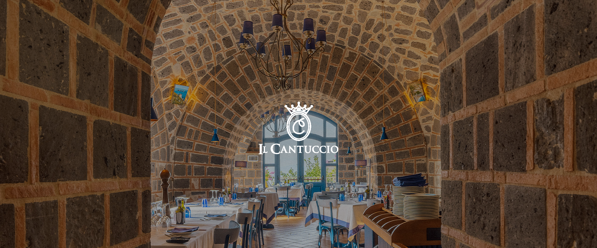 Interior view of a restaurant with arched brick ceiling, set tables with white tablecloths, napkins, and glassware, large window at the back, and a chandelier hanging from the ceiling.