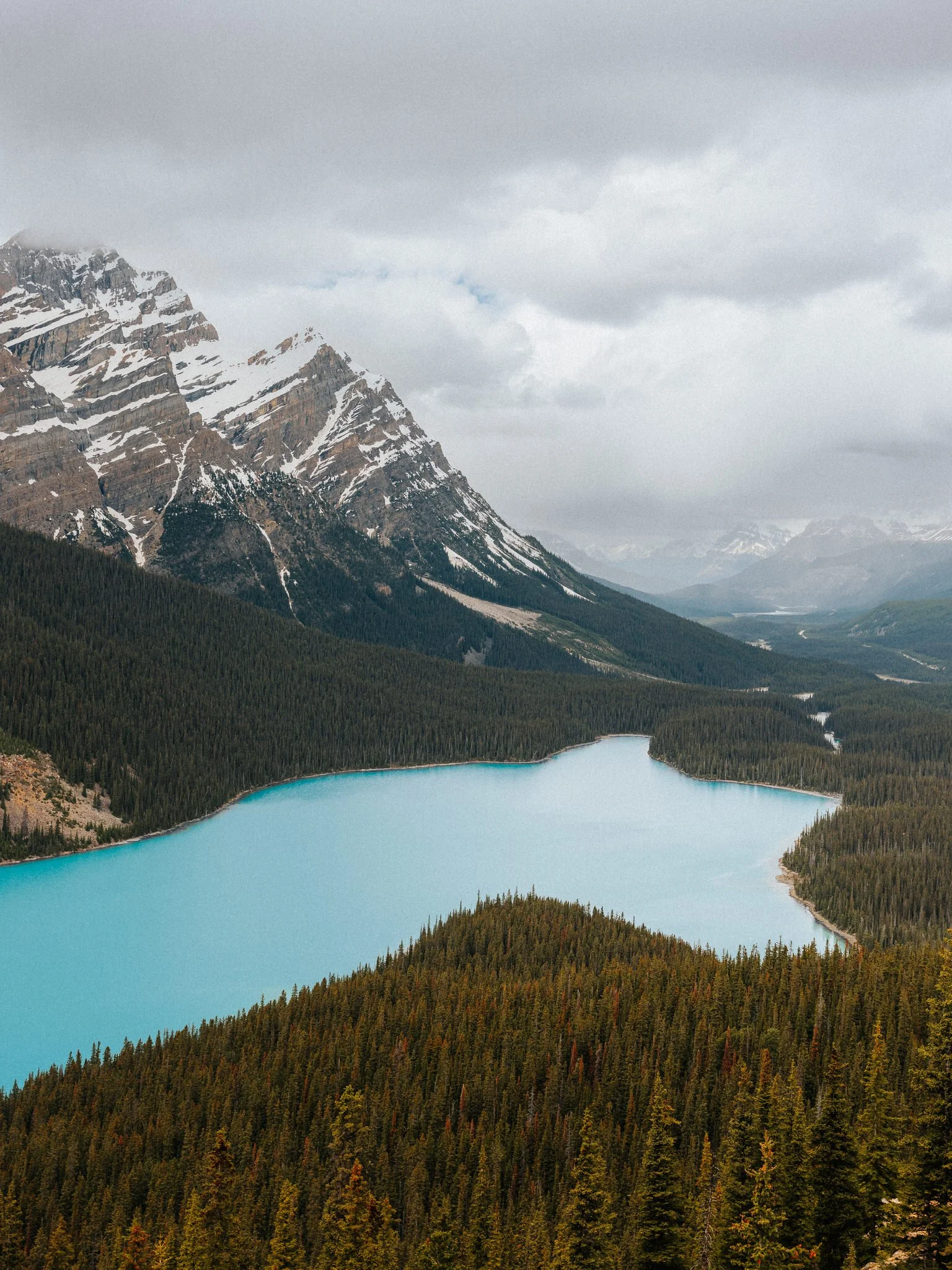 Scenic view of a turquoise lake surrounded by dense evergreen forest with snow-capped mountains and cloudy sky in the background.