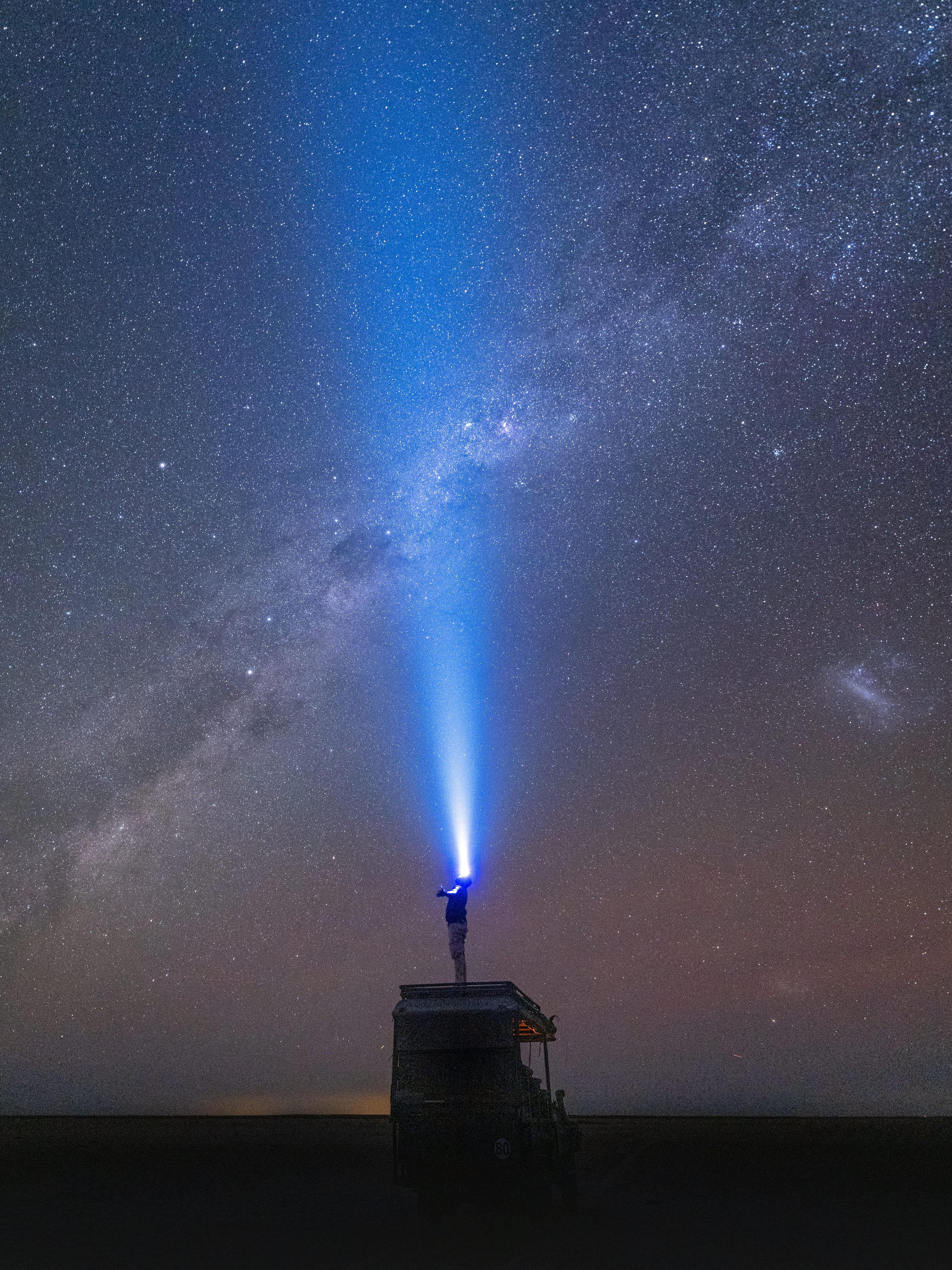 Person standing on a platform with a flashlight directing a beam of light into the night sky filled with stars and the Milky Way galaxy.