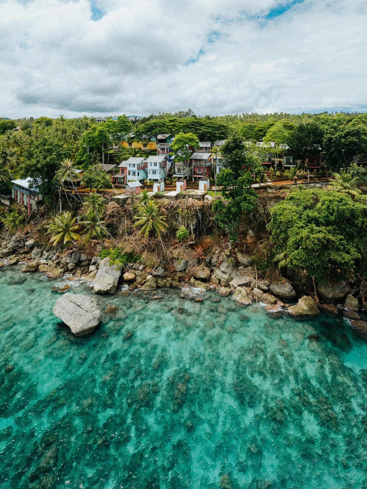 Aerial view of a coastal town with colorful houses on a cliff, surrounded by lush green trees and palm trees, above clear turquoise water with rocks.