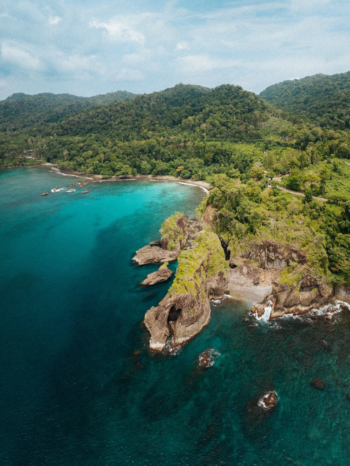 Aerial view of a tropical coastline with green hills, rocky cliffs, and clear blue ocean water.