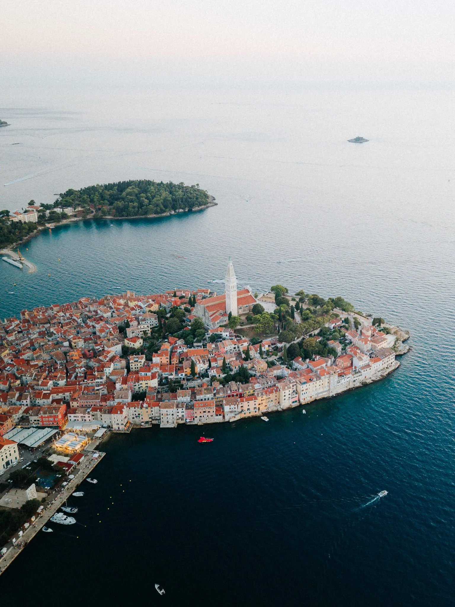 Aerial view of historic Croatian town on a peninsula, with red-tiled roofs, church steeple, and boats on dark blue sea.