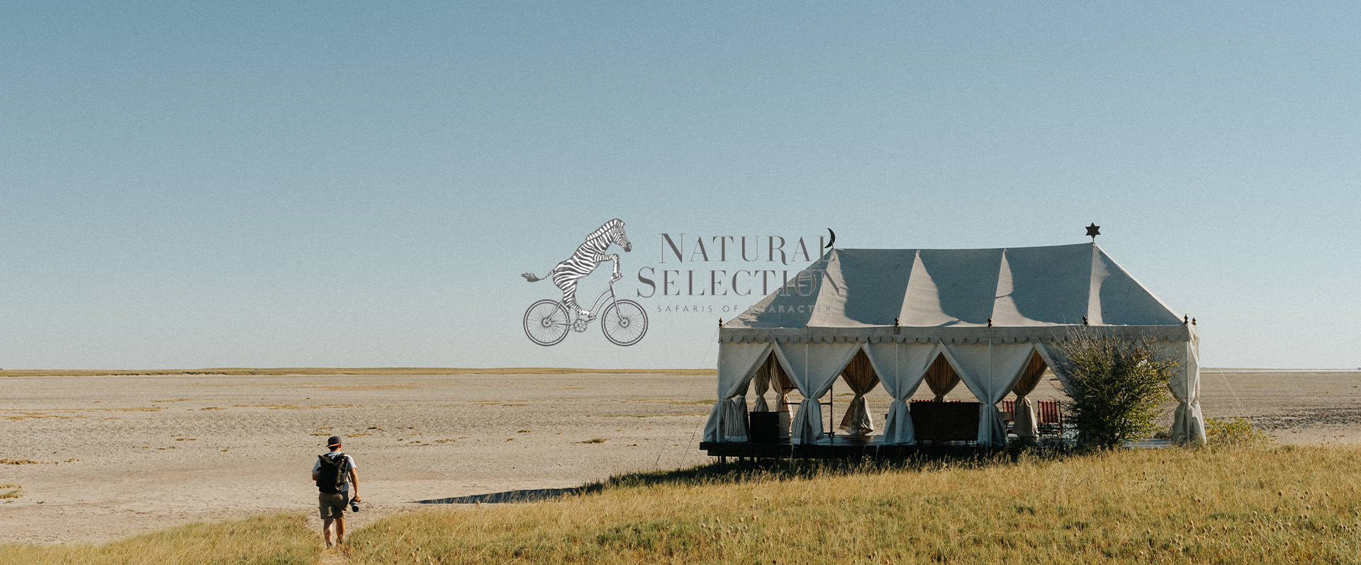A person with a backpack walking towards a large white tent in an open grassy and sandy landscape under a clear blue sky, with a logo in the sky that says "Natural Selection Safaris of Character" featuring a zebra riding a bicycle.