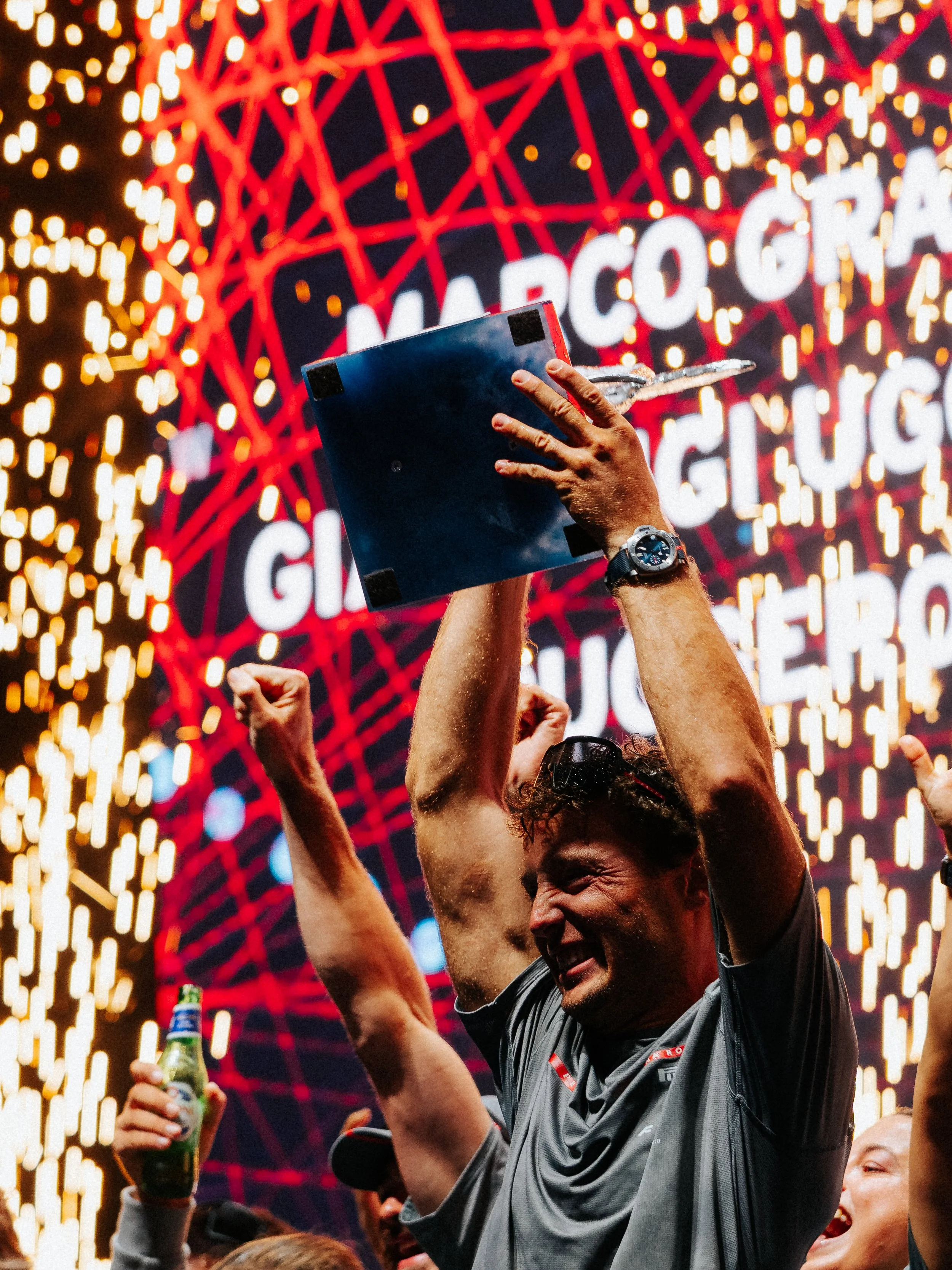A man celebrating with arms raised, holding a bottle of beer, at a lively event with other people, bright lights, and a large sign that says 'MARIO GRA'}