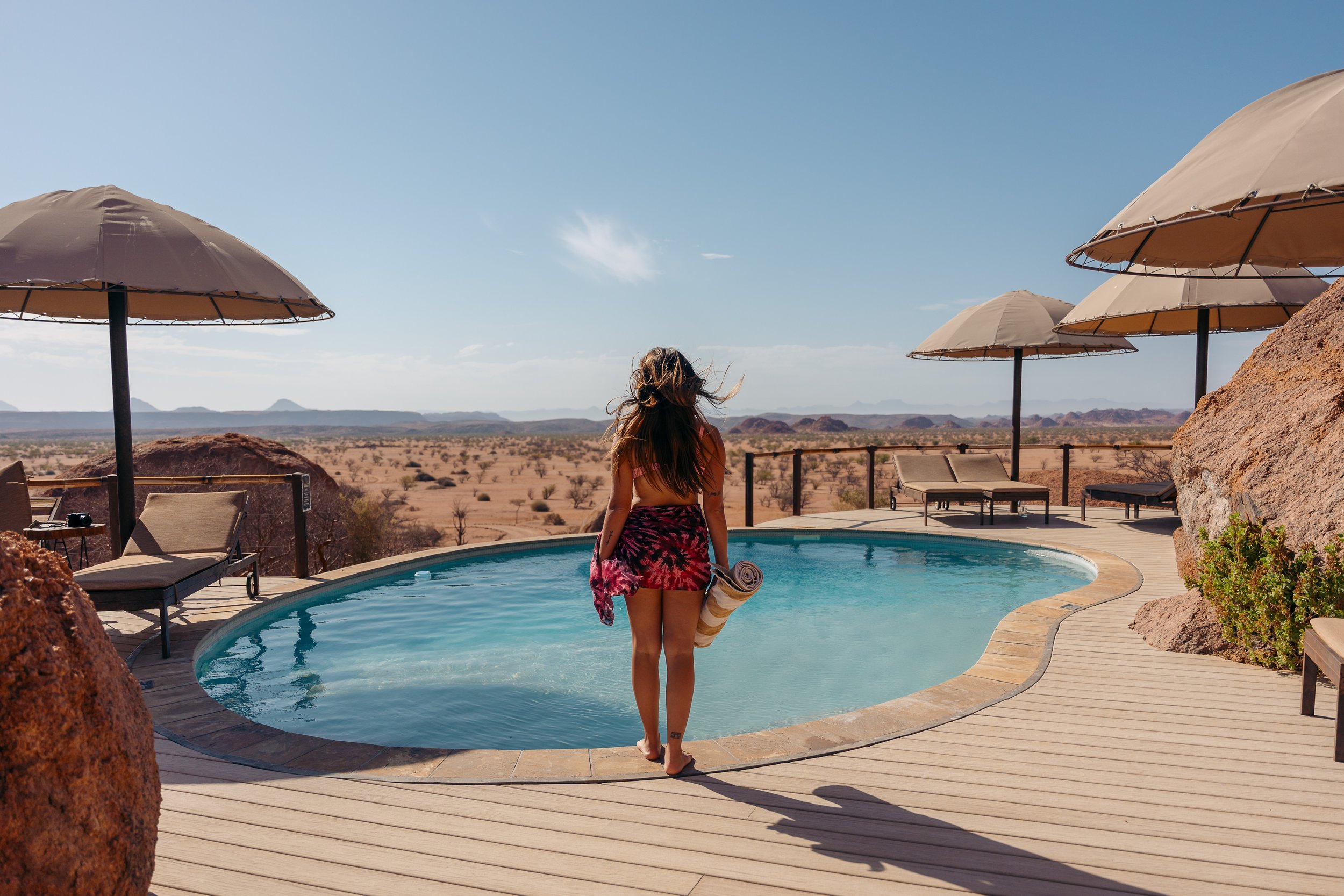 A woman walking by a swimming pool in a desert landscape with umbrellas and lounge chairs