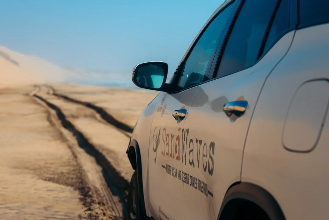 Close-up of a white car traveling on a desert road with sand dunes in the background, and a logo on the car that reads #SandHavens.