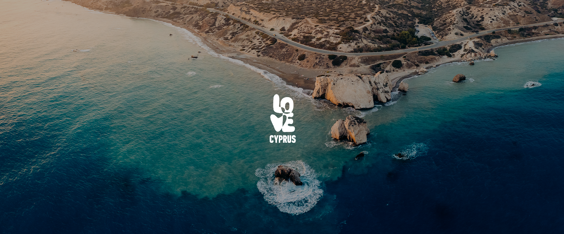 Aerial view of a coastal area in Cyprus with rocky shoreline, sandy beach, turquoise water, and rugged hills in the background, featuring the Love Cyprus logo in the center.