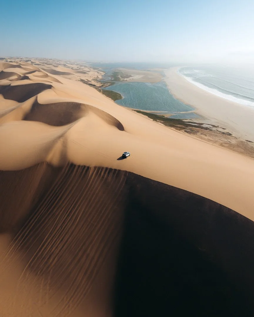 Aerial view of sand dunes with a vehicle on the sand near a water body, possibly a river or lake, with an ocean shoreline in the background.