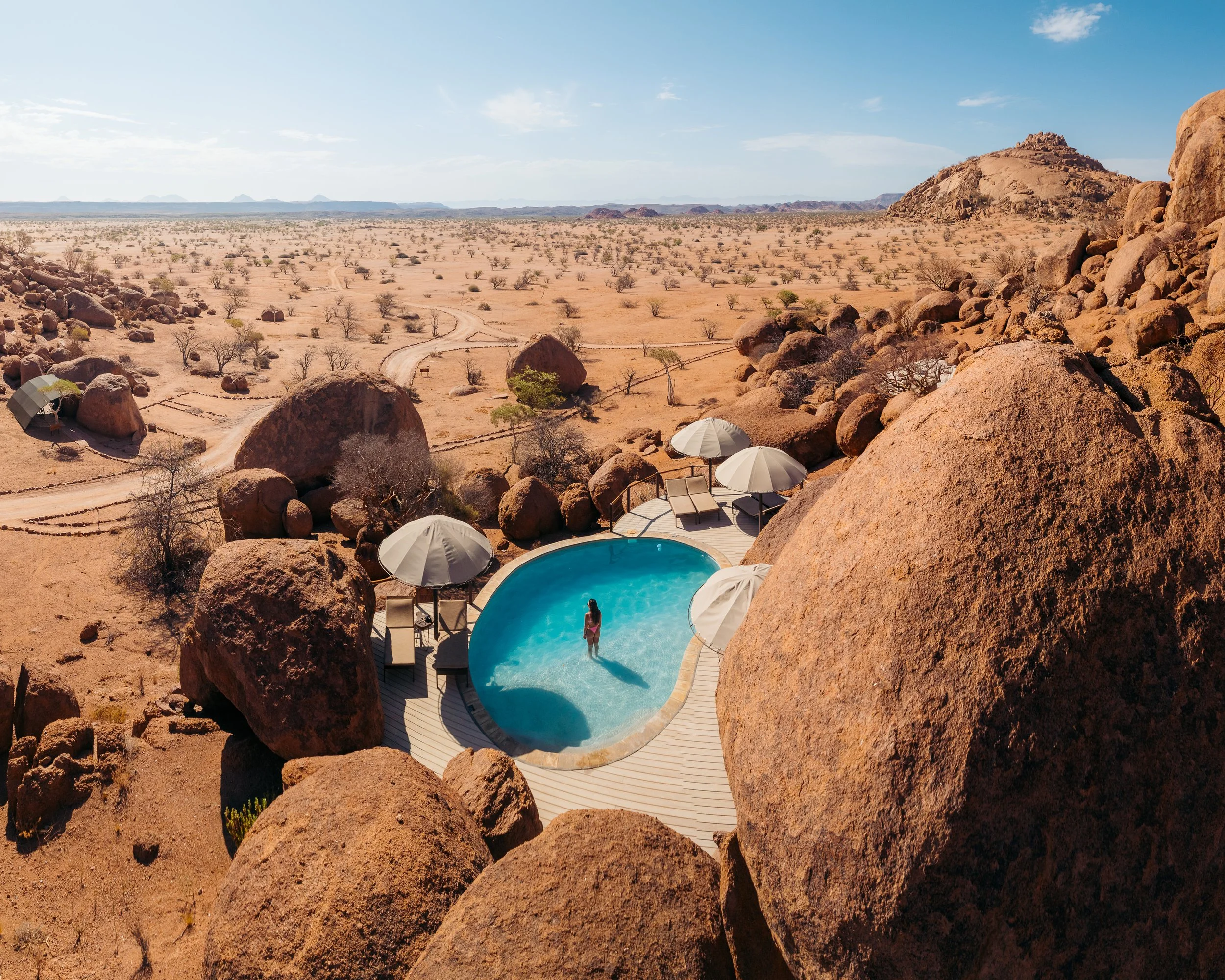A woman standing in a small, kidney-shaped swimming pool surrounded by large rocks, with desert landscape and sparse bushes in the background.