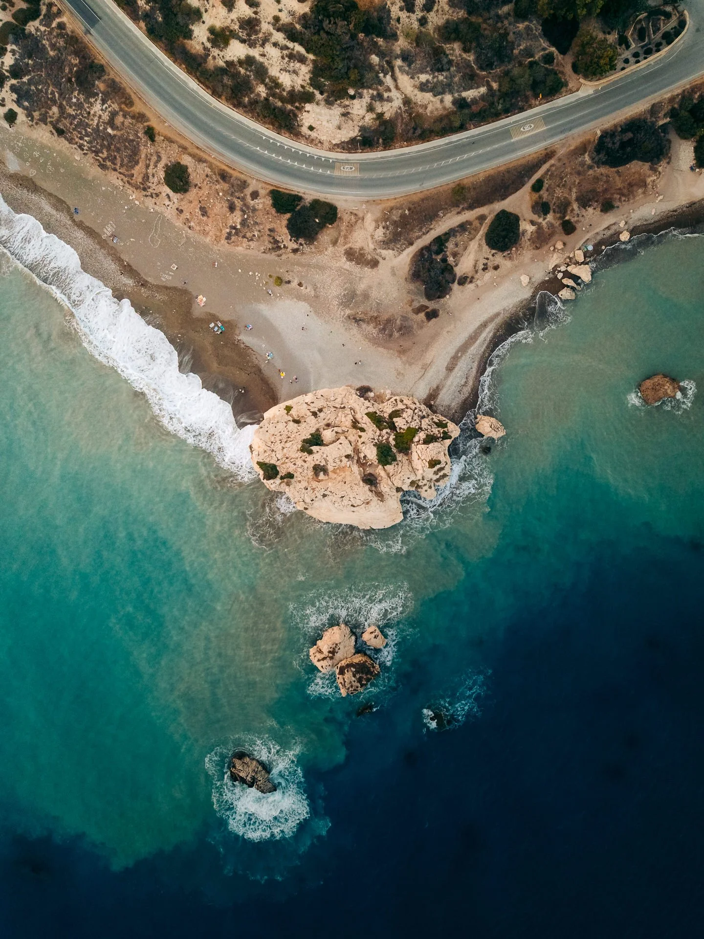 An aerial view of a coastal area showing a winding road parallel to the shoreline, a beach with small umbrellas and people, large rocks in the water, and turquoise ocean waves crashing on the beach.