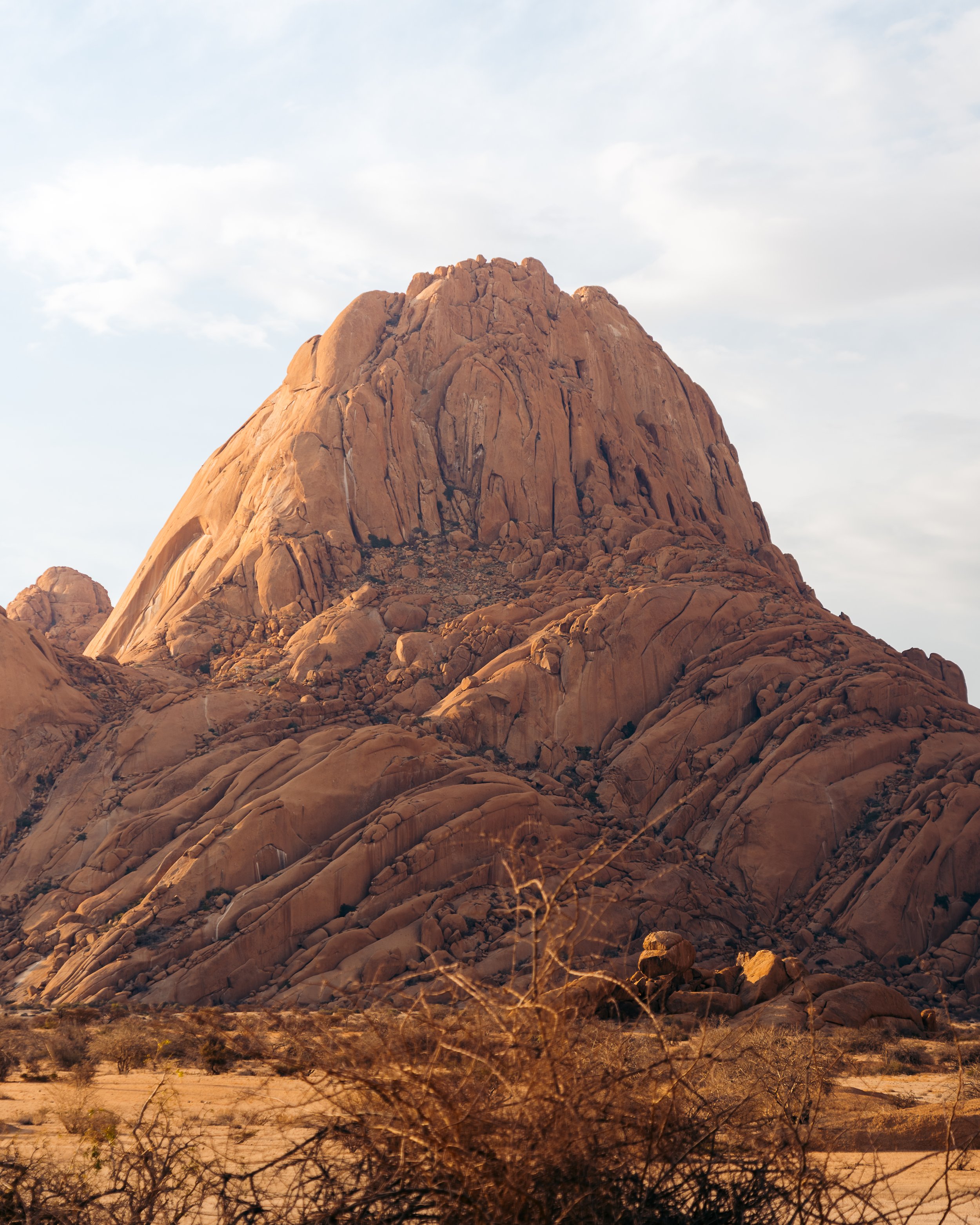 A large, rugged, orange-brown mountain rising in a desert landscape with sparse desert plants in the foreground and a partly cloudy sky.