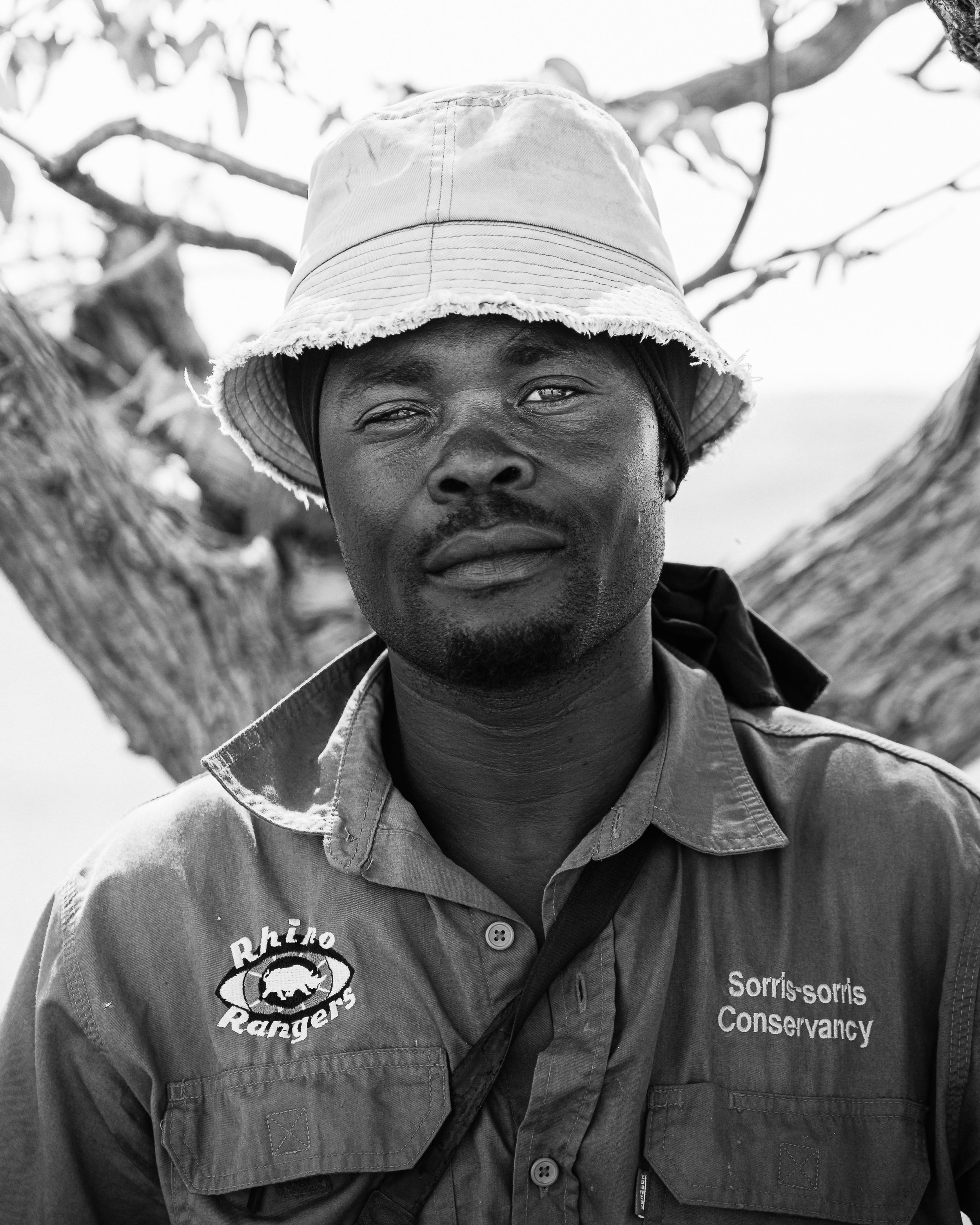 A man wearing a hat and outdoor clothing with logos for 'Rhino Rangers' and 'Sorris-soris Conservancy' stands outdoors in front of a tree.