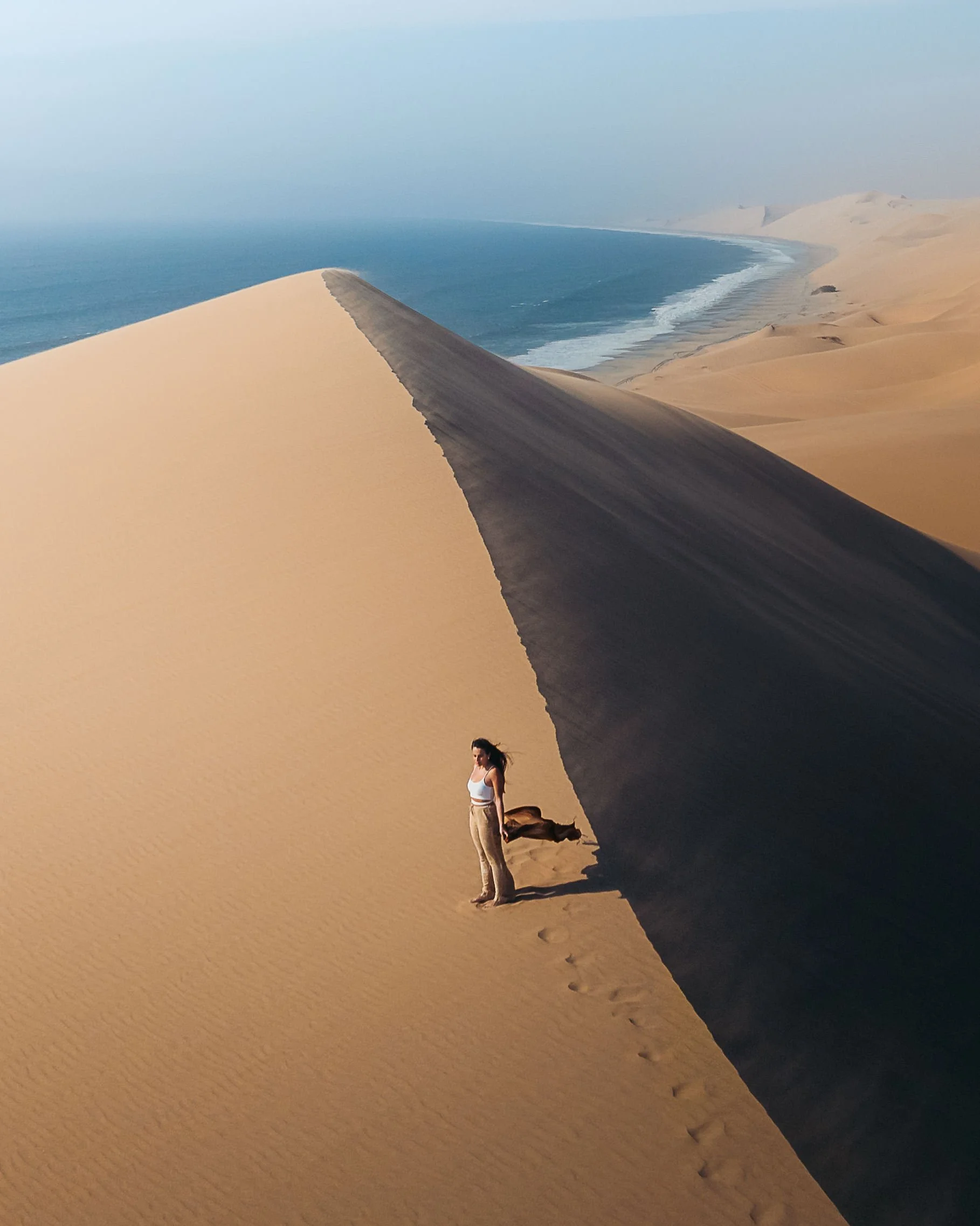 A woman stands on a sand dune with one side illuminated by sunlight and the other side in shadow, near an ocean coastline with waves.