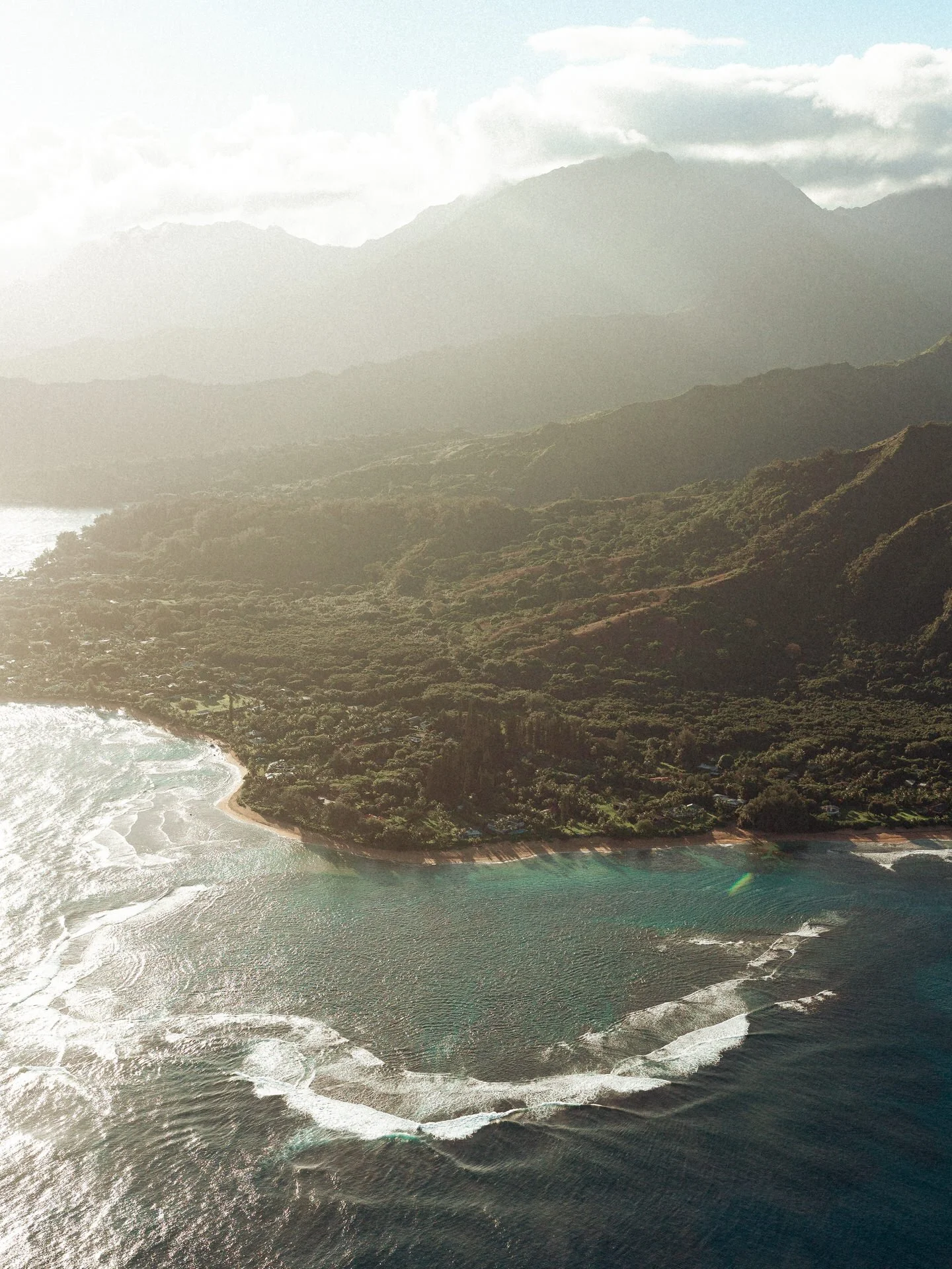 Aerial view of a coastline with clear water, waves, and lush green hills with mountains in the background.