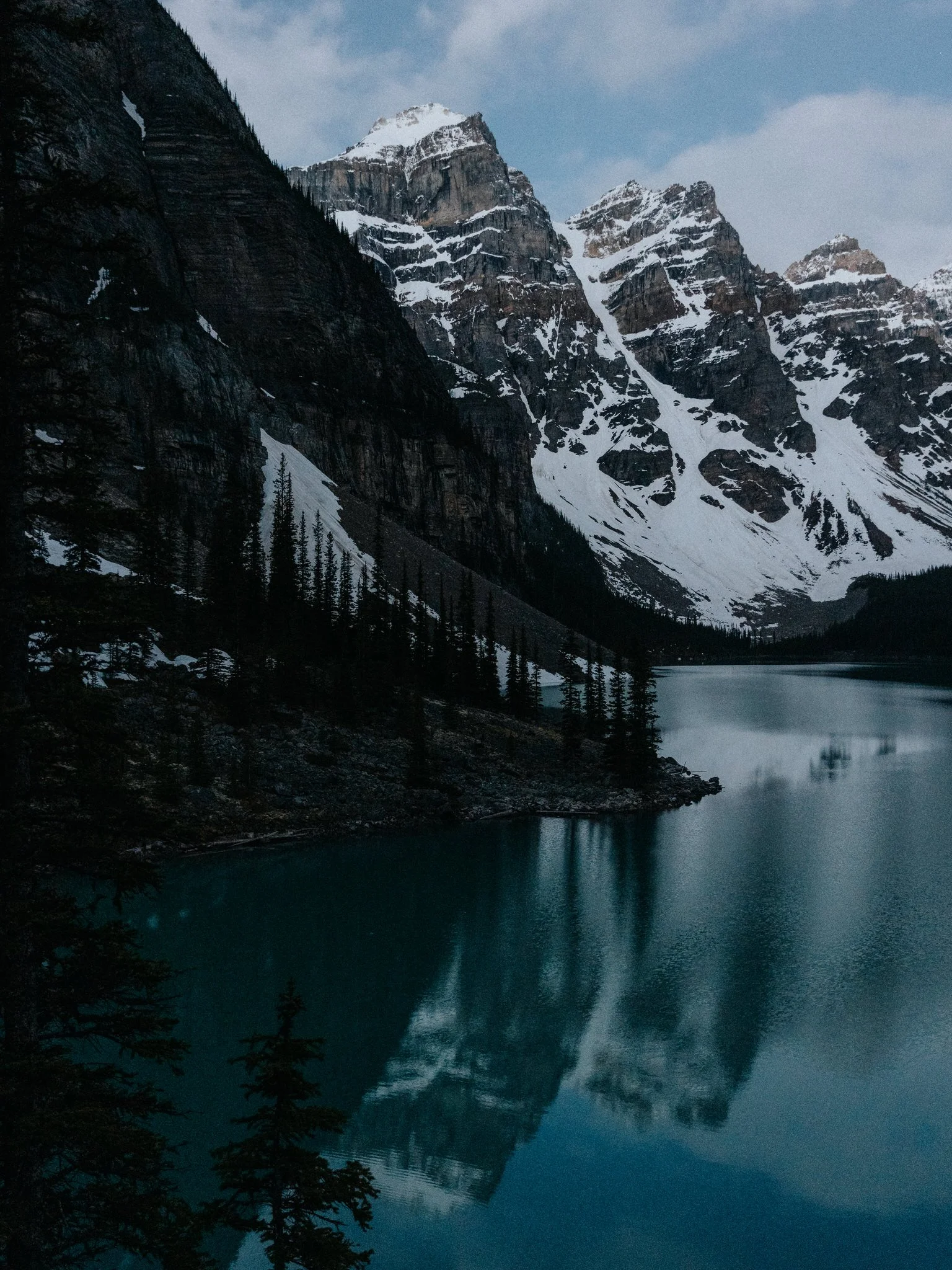 Snow-capped mountains reflected in a calm lake, with dark pine trees along the shoreline under a blue sky.
