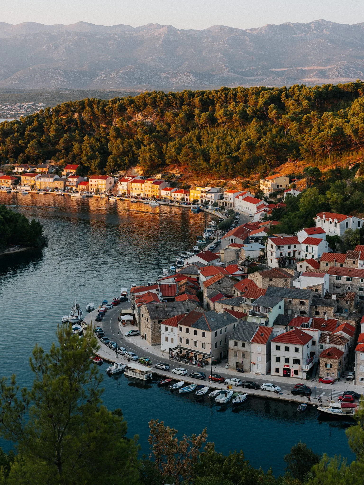 A picturesque coastal town with white buildings and red rooftops along a harbor, surrounded by lush green trees and a mountain range in the background at sunset.
