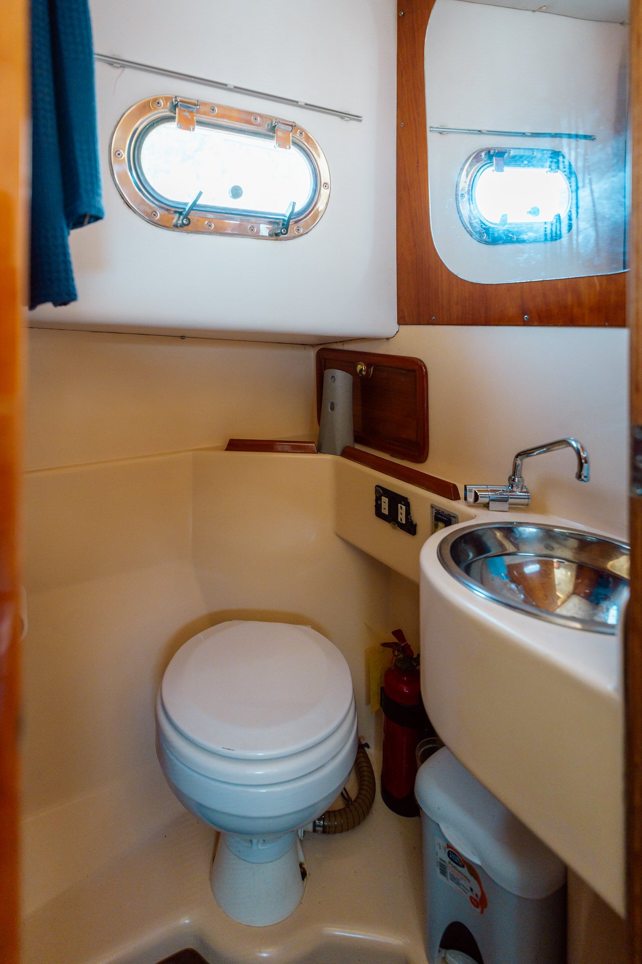 Small boat bathroom with oval porthole windows, a toilet, a small sink, and a mirror, with wood accents and a fire extinguisher nearby.