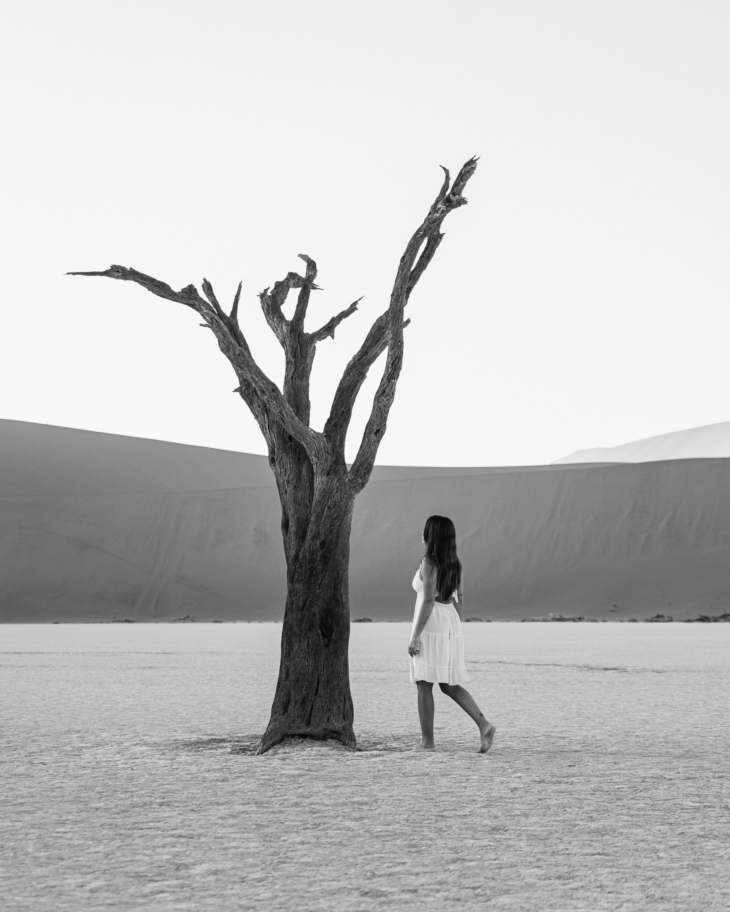 A woman in a white dress standing barefoot in a desert next to a dead tree, with sand dunes in the background, in black and white.