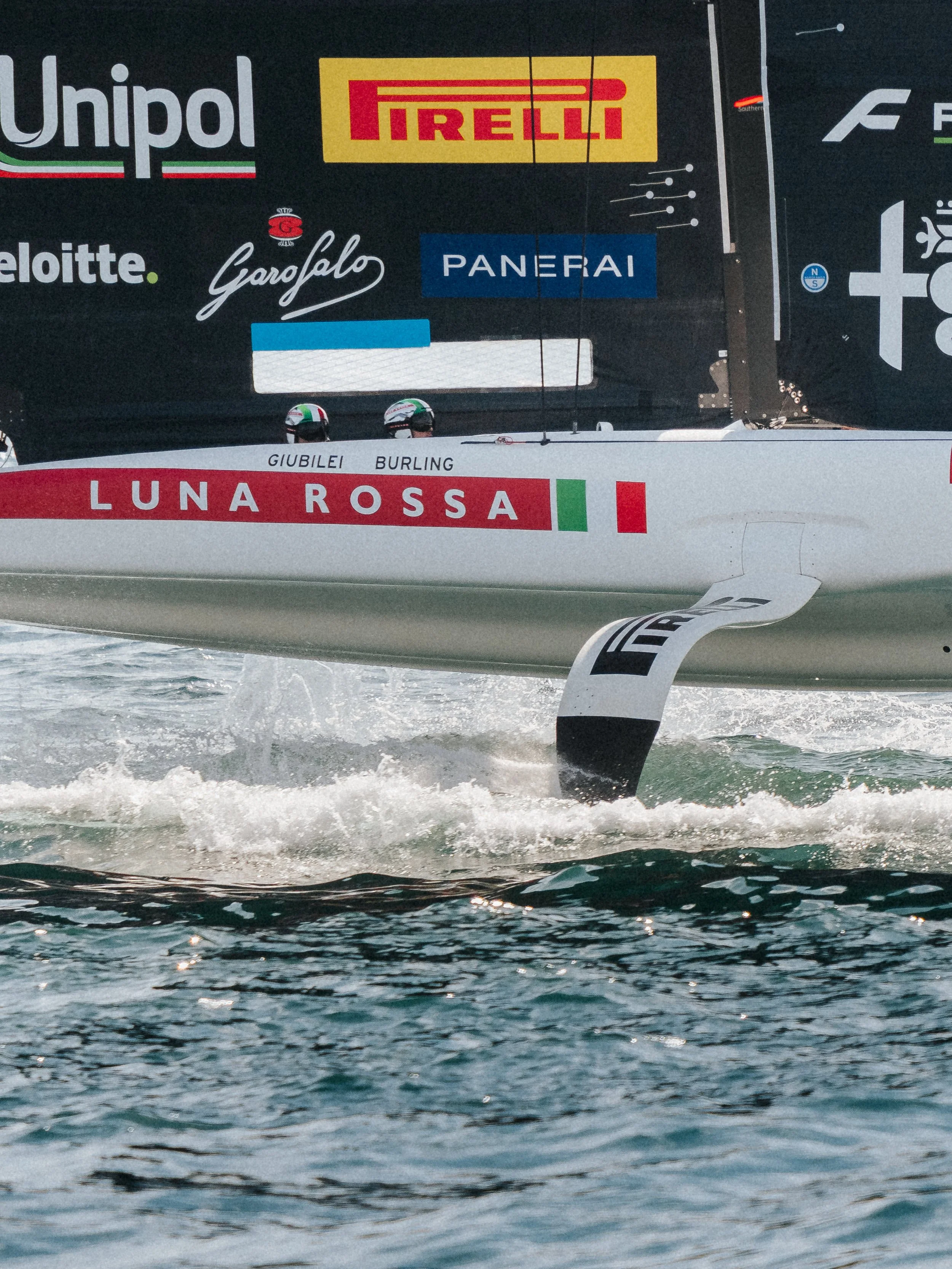 Close-up of a sailboat moving through the water with sponsors' logos on its sail, including Unipol, Pirelli, Deloitte, Garofalo, Panerai, and Luna Rossa, with two sailors visible at the bottom wearing helmets.