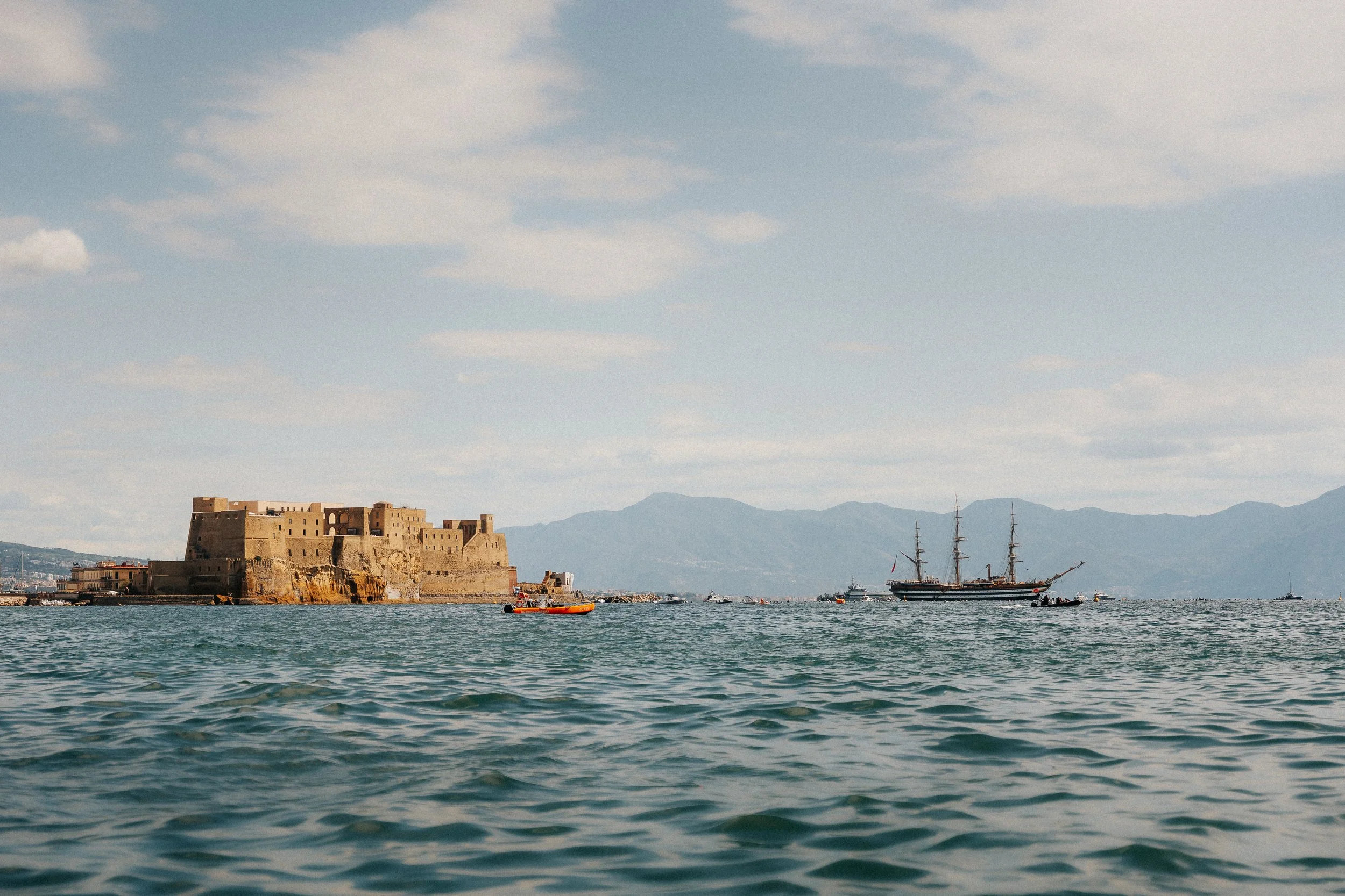 A harbor scene with a historic stone castle on the shoreline, several sailboats and a sailing ship in the water, mountains in the background, and a partly cloudy sky.