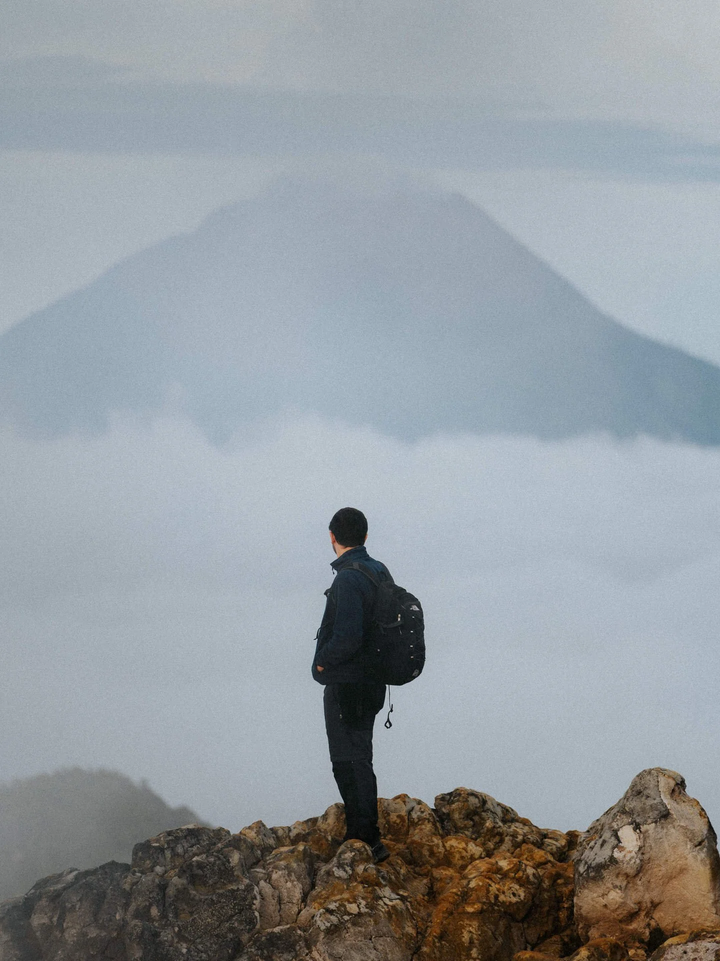 Man with a backpack standing on rocky terrain, looking at distant mountain and cloud layers.