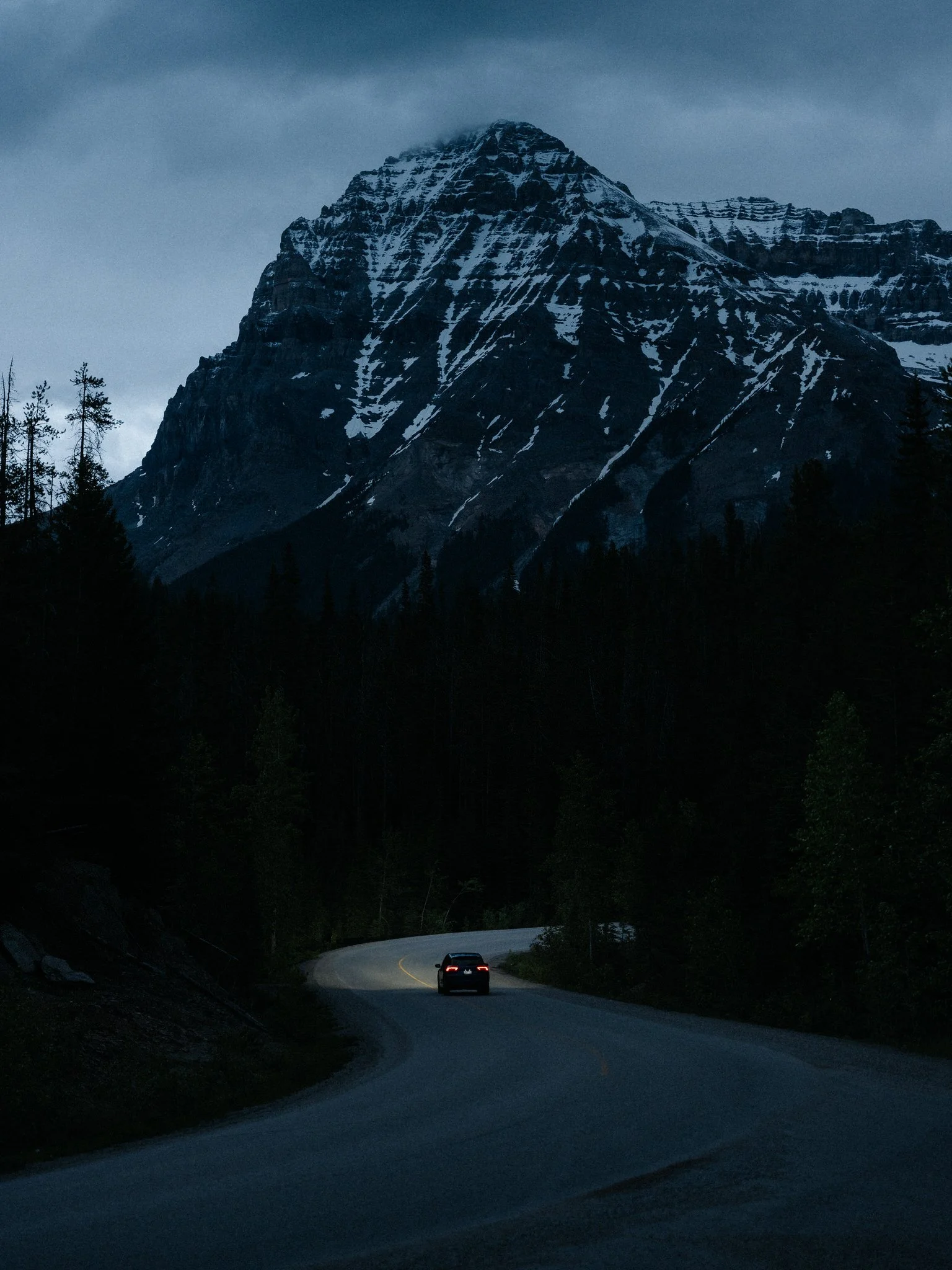 A dark mountain landscape with a winding road and a small car driving away, with snow-capped peaks and tall trees.
