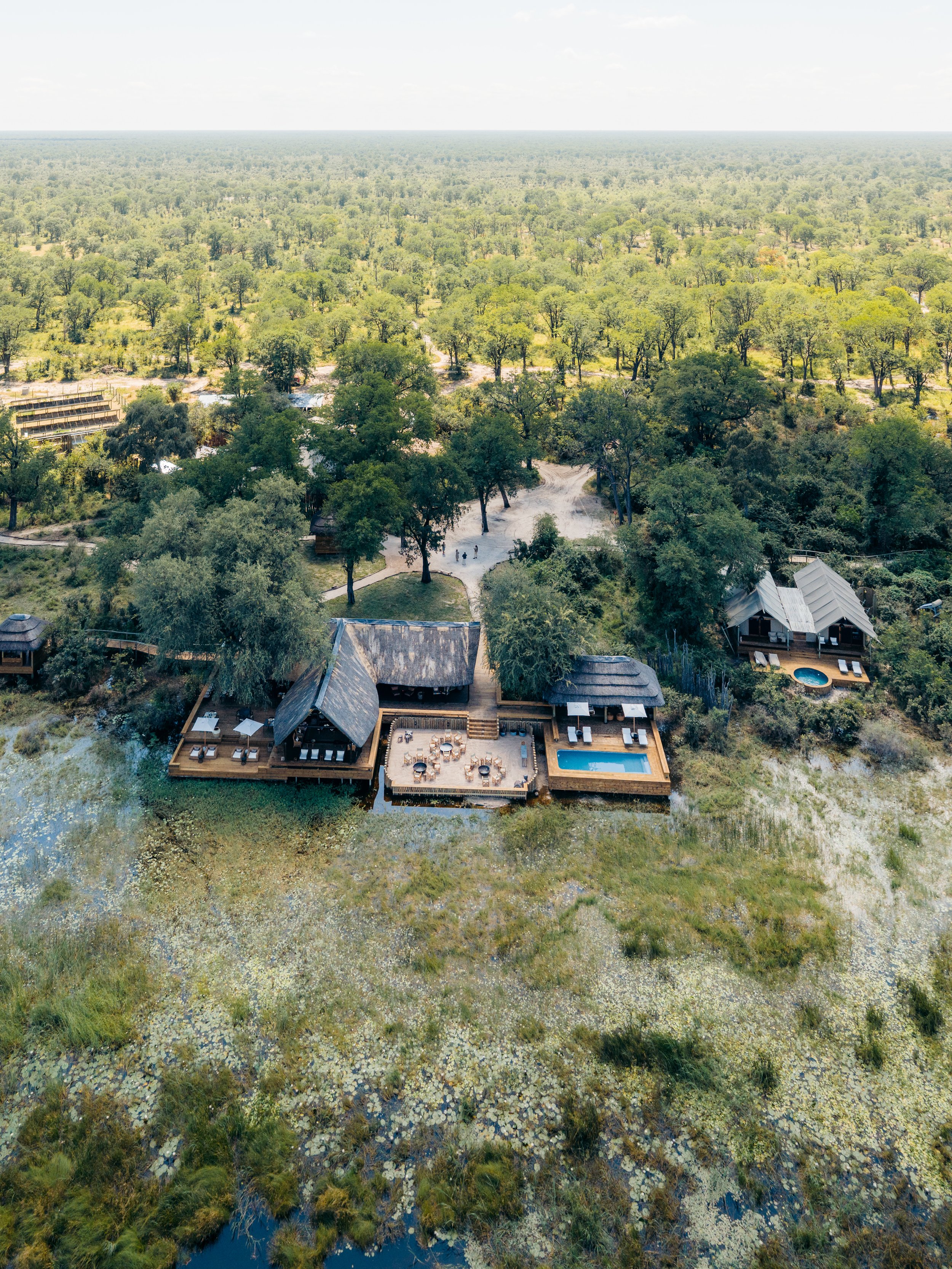 Aerial view of a natural landscape with dense green trees, a few structures, a body of water, and a recreational area with a pool and outdoor seating.